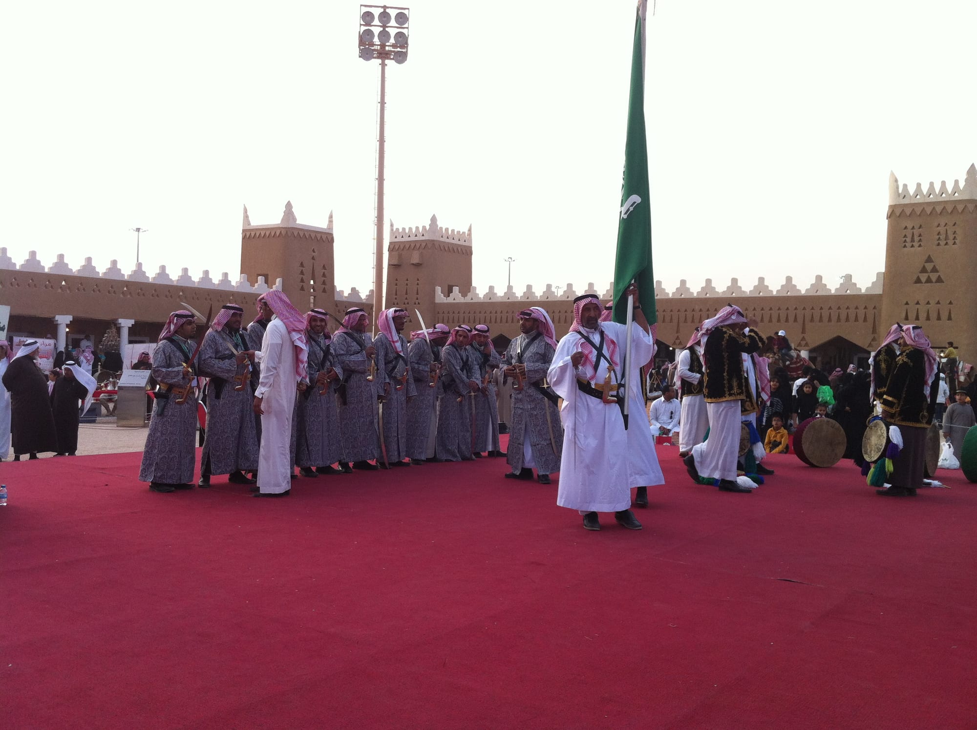 Rows of men in grey robes and red-checked headscarves holding swords perform the ardah on a red carpet, while a man in a white thobe raises the green Saudi flag and drummers stand to one side before crenellated mud-brick walls