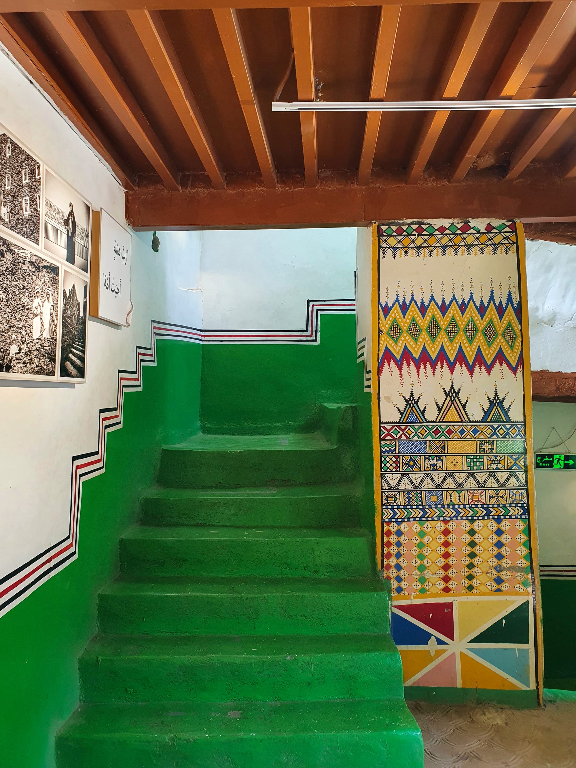 A green-painted stone staircase beside a white wall edged with stepped black, red and white lines, with a pillar covered in bright geometric painted bands of diamonds, triangles and zigzags
