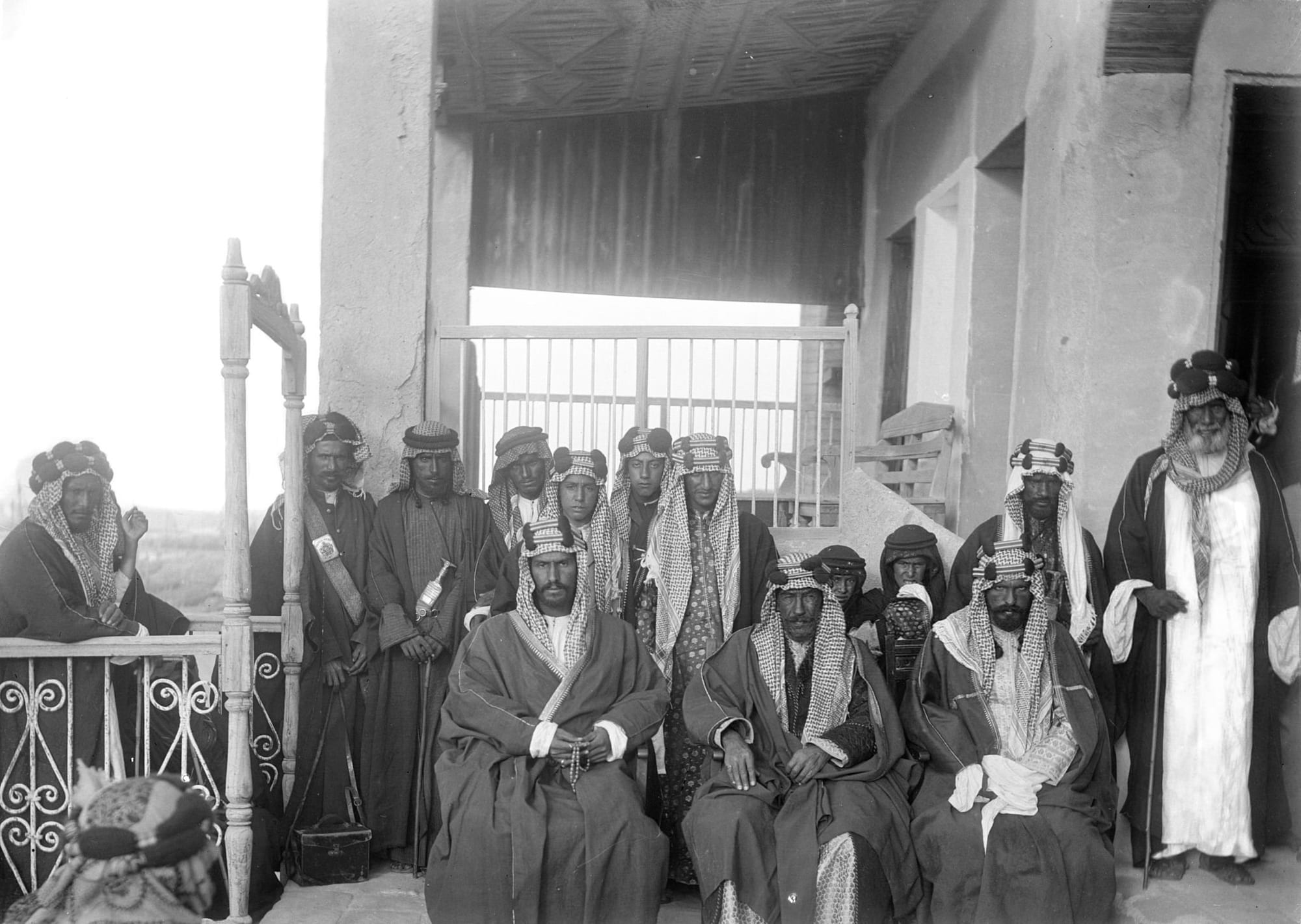 A black-and-white photograph from 1910 of three men in robes and checked headcloths seated on a veranda, with a row of attendants standing behind them