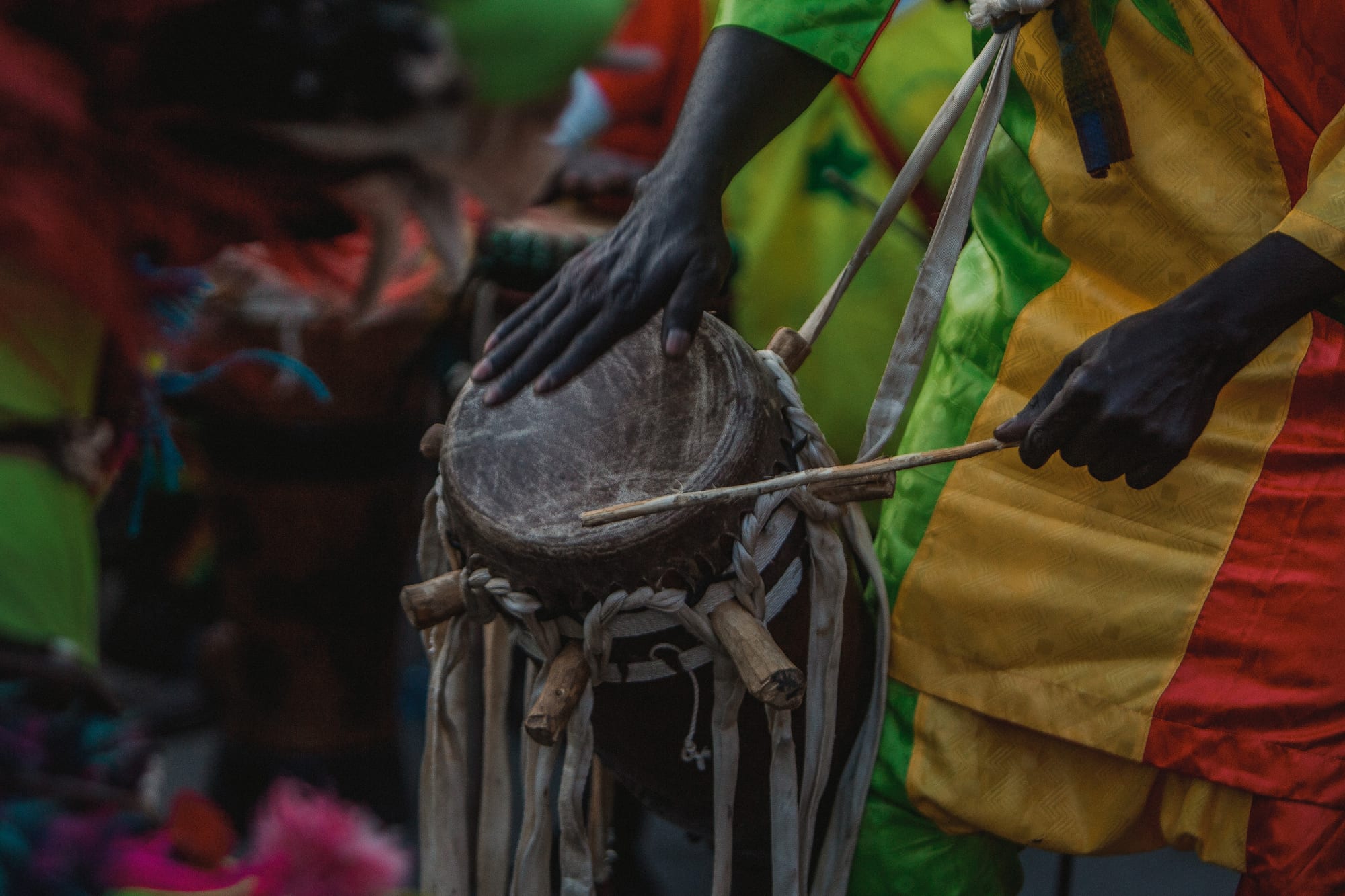 A close view of a small sabar drum, bound with cord and wooden pegs, played by a drummer in a green, yellow and red robe using one hand and a thin stick