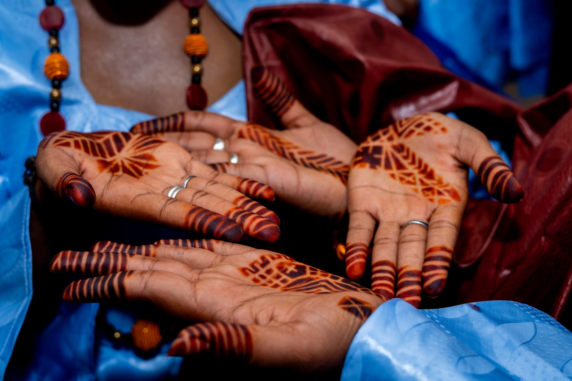 Several women's hands held together with palms up, decorated with bold dark red henna patterns on the palms and fingertips, above pale blue and maroon robes