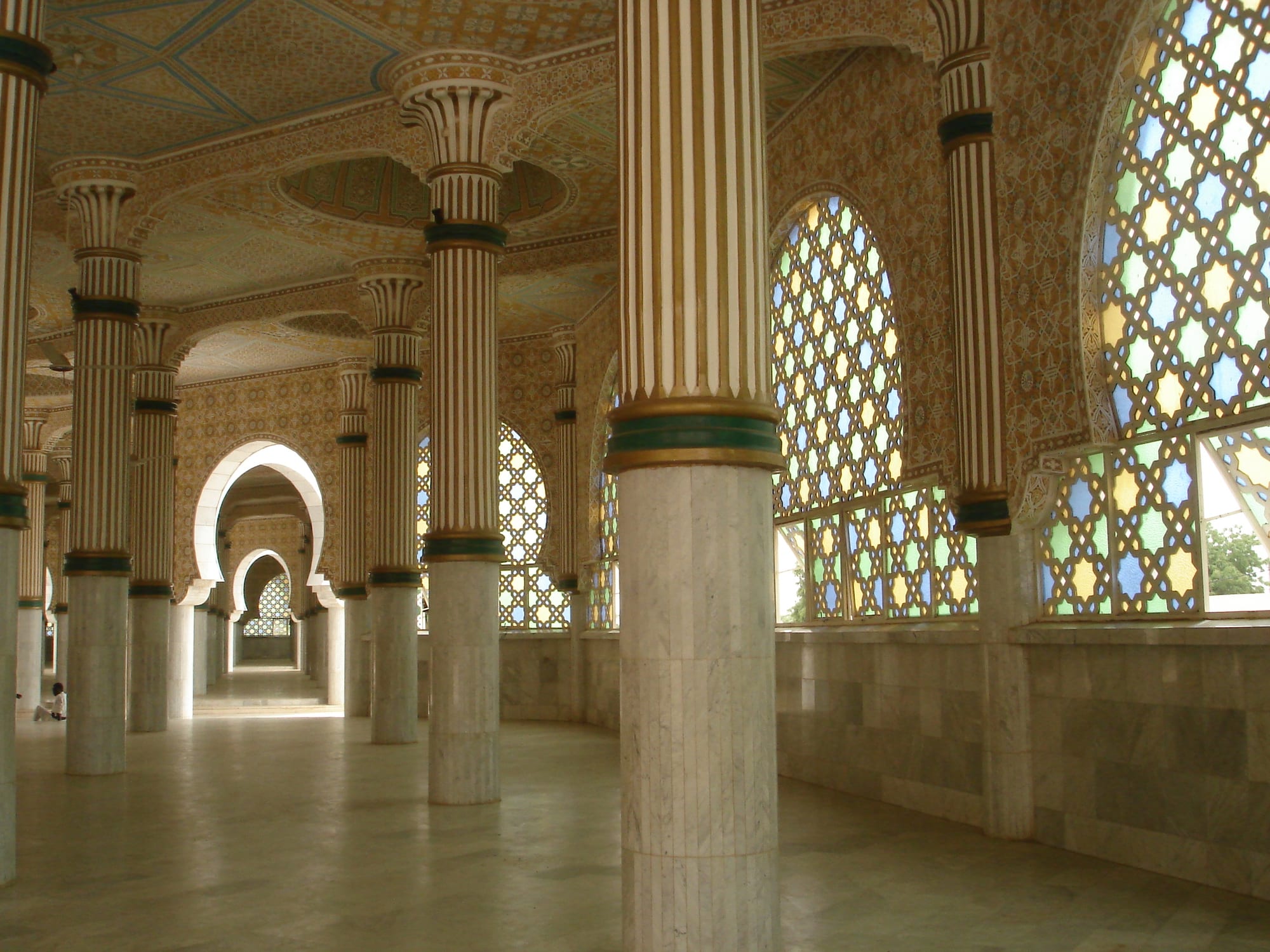 A long marble colonnade inside a mosque, with fluted columns, a painted ceiling and tall arched windows of coloured geometric glass