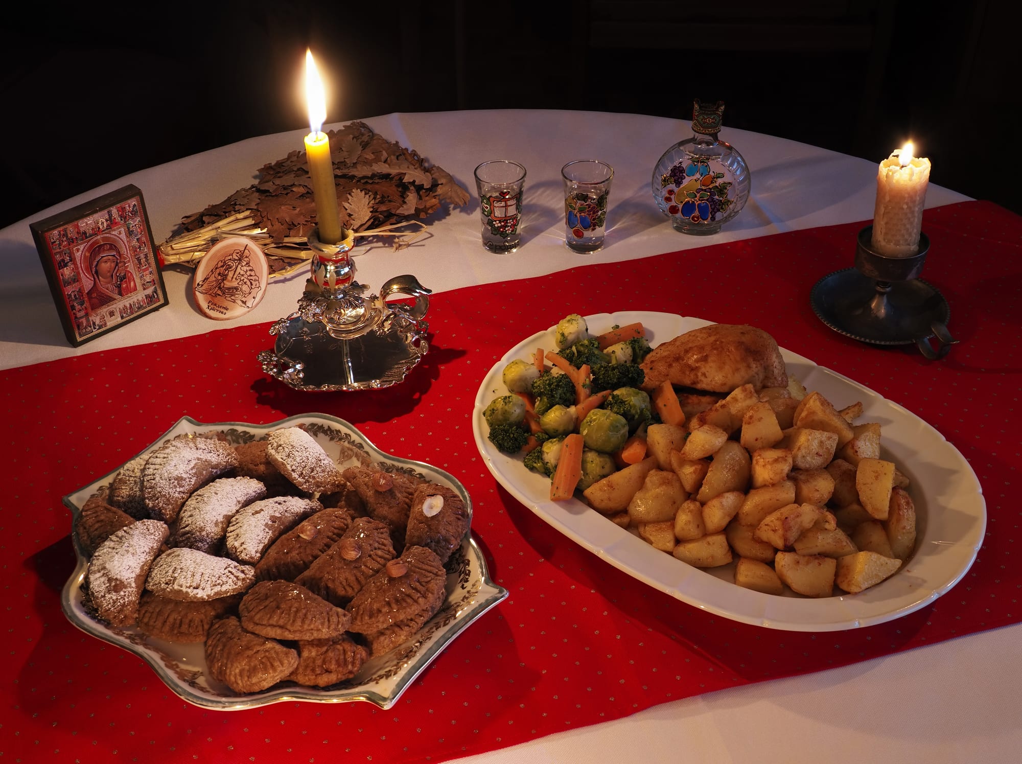 A candlelit table with a red runner, an icon of the Virgin, a bundle of dried oak leaves, small glasses, a plate of sugar-dusted biscuits and a platter of chicken, potatoes and vegetables