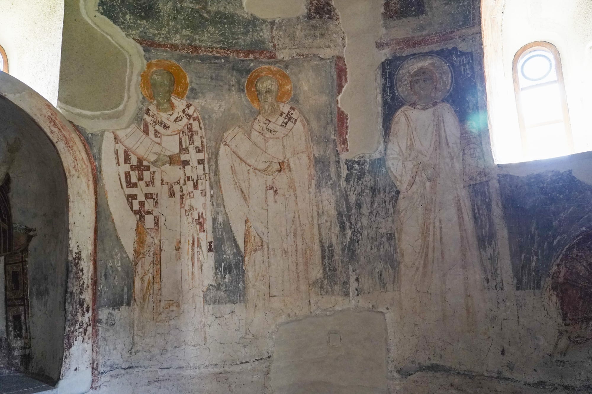 Faded medieval frescoes of haloed saints in patterned robes painted on the curved wall of a church, lit by a small arched window