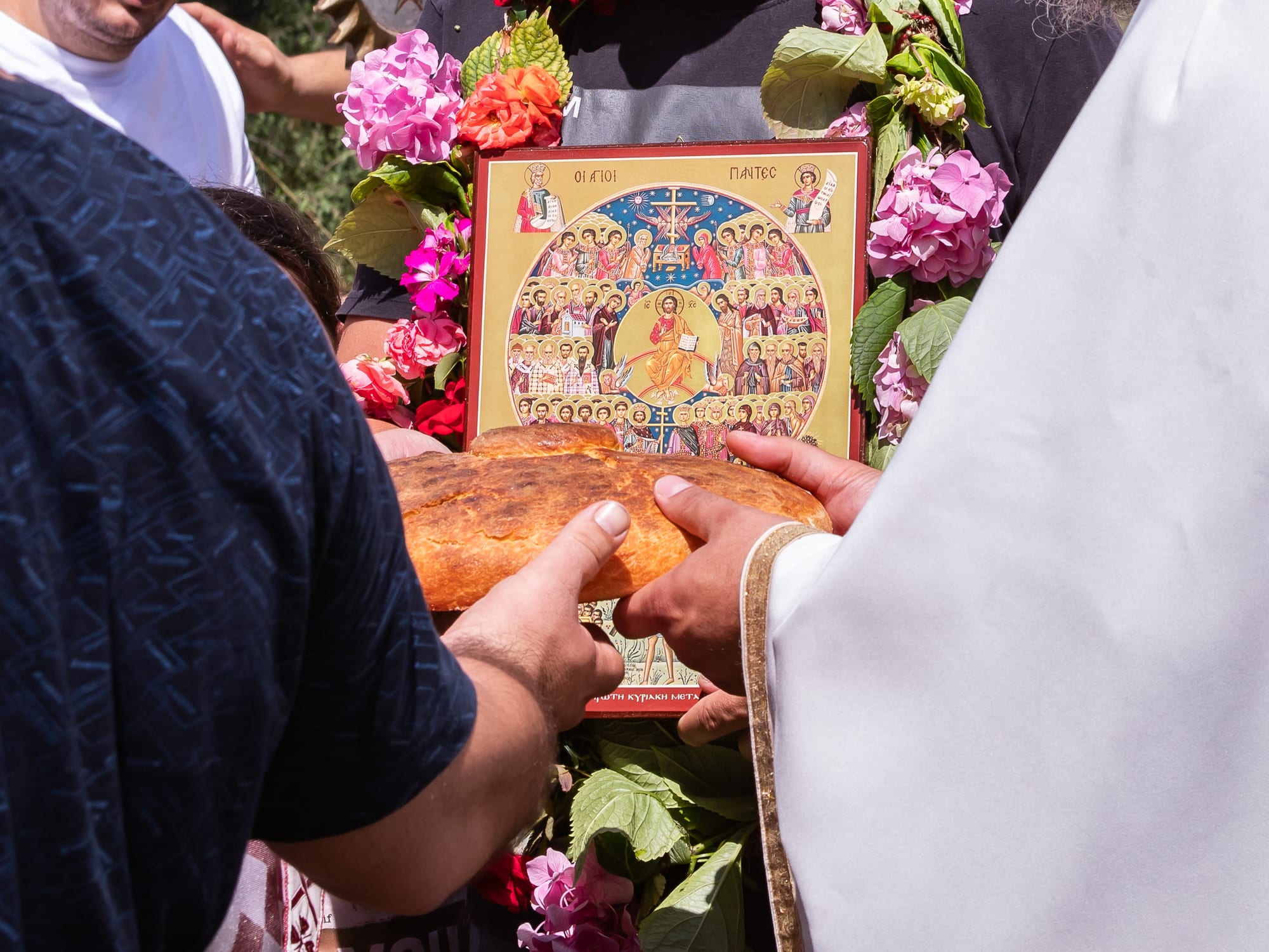 Two pairs of hands, one belonging to a priest in white vestments, hold a round loaf of bread in front of an icon of the saints framed with pink and red flowers
