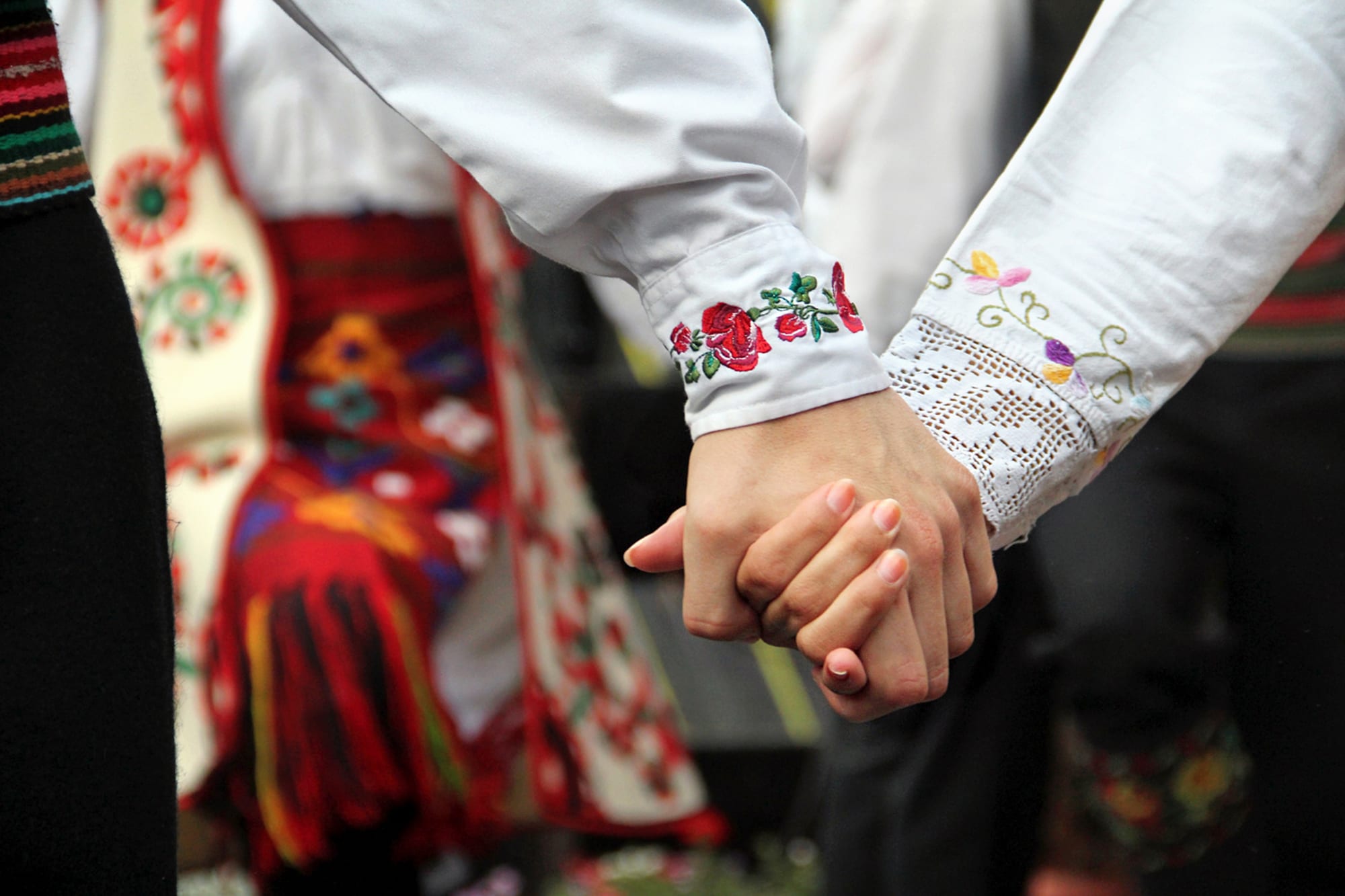 Close view of two dancers' joined hands, their white sleeves embroidered with red roses and finished with lace, against brightly patterned folk costume