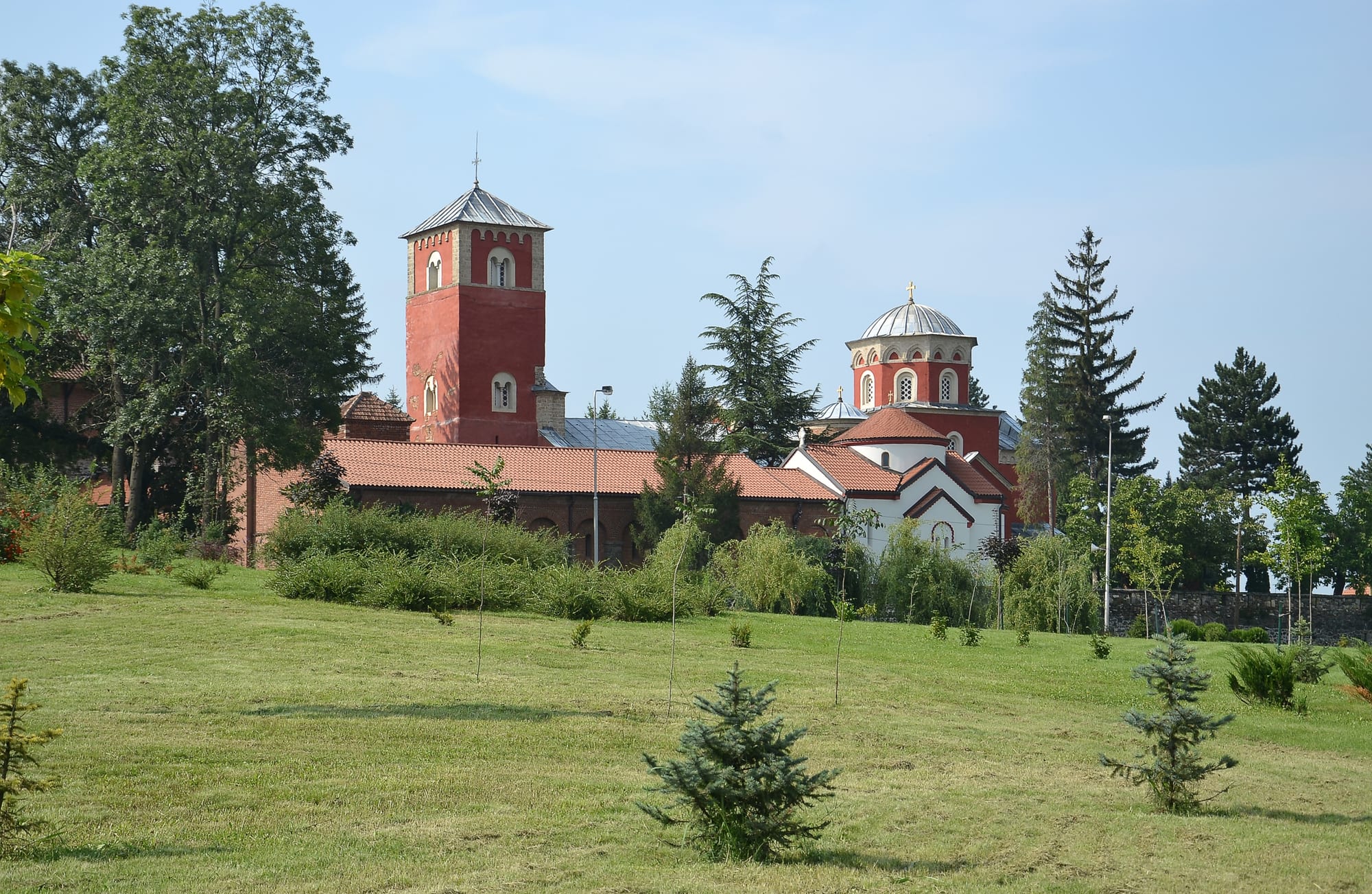 The red-painted bell tower and domed church of Žiča monastery among trees, seen across a lawn