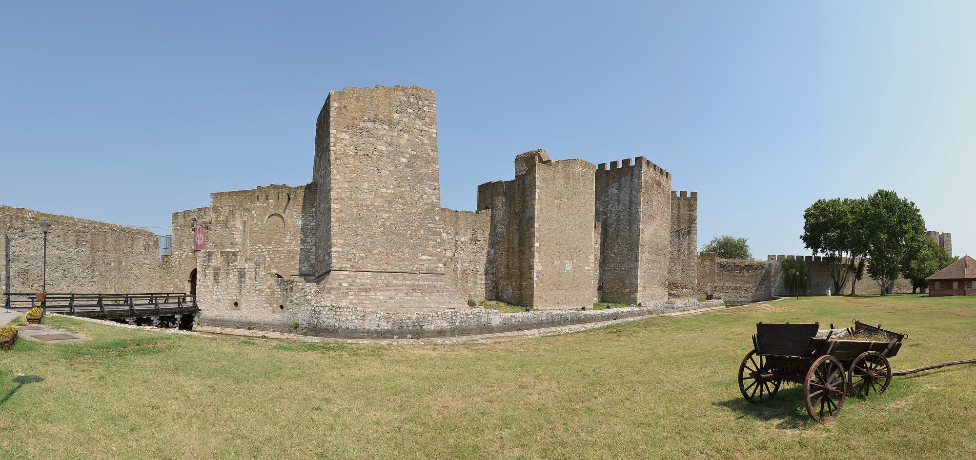 The square stone towers and crenellated walls of Smederevo fortress rising above a grass field, with a wooden cart in the foreground