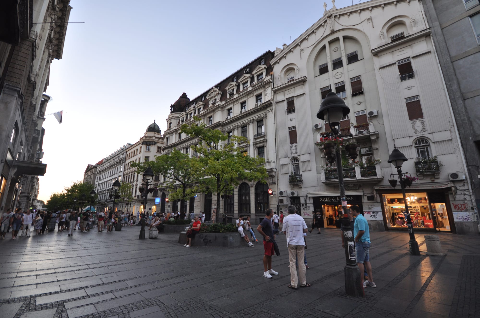A crowded pedestrian shopping street in Belgrade at dusk, with people standing and sitting around benches and shopfronts lit up