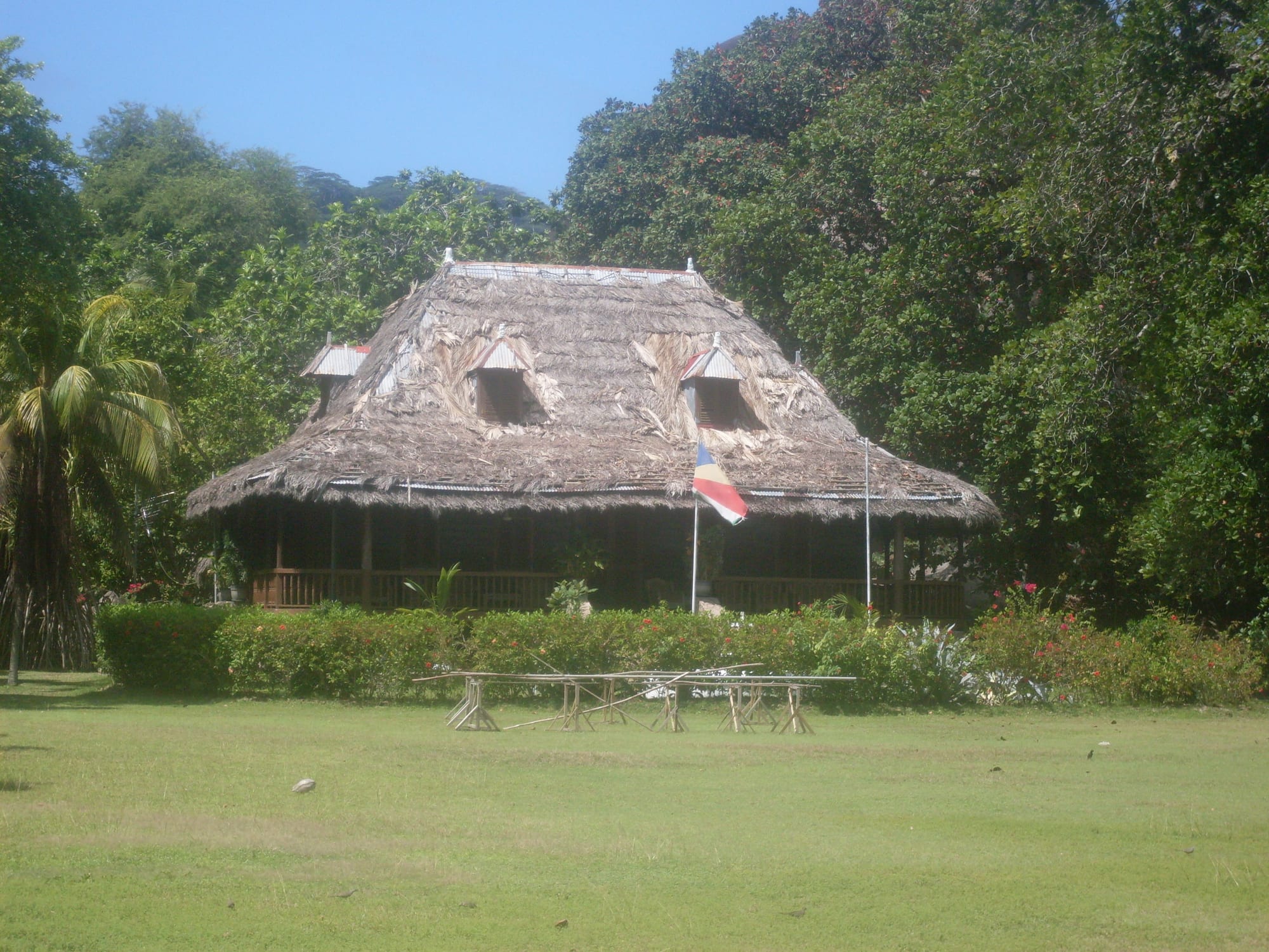 A large plantation house with a steep thatched roof, dormer windows and a shaded veranda, set on a lawn in front of dense green trees, with a Seychelles flag on a pole