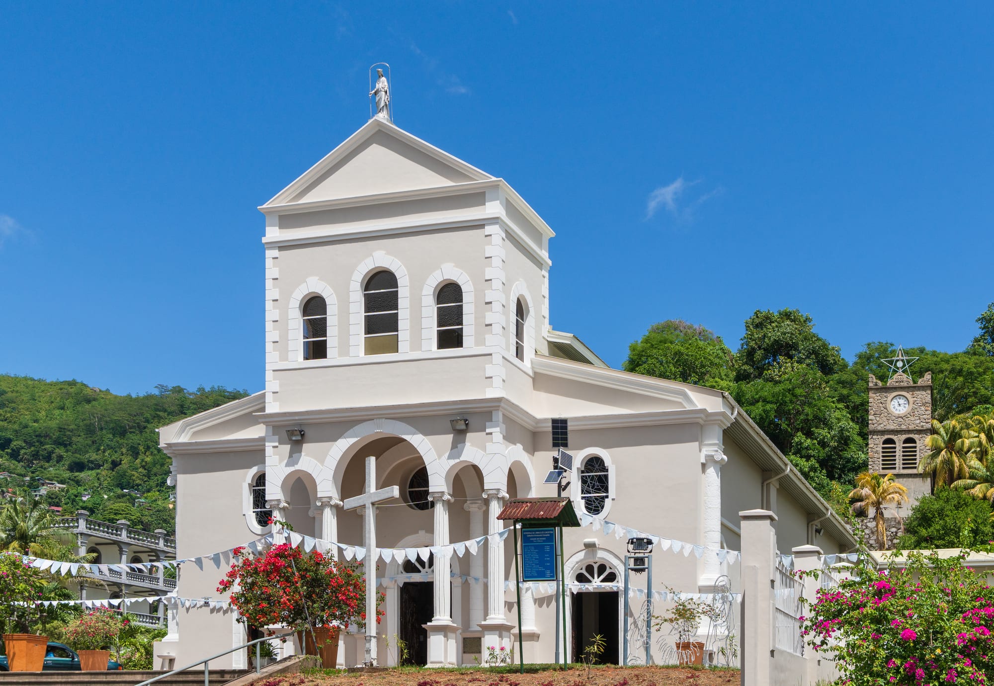 A white church with arched windows and a statue on its gable, strung with white bunting and framed by bougainvillea, with a stone clock tower and green hills behind