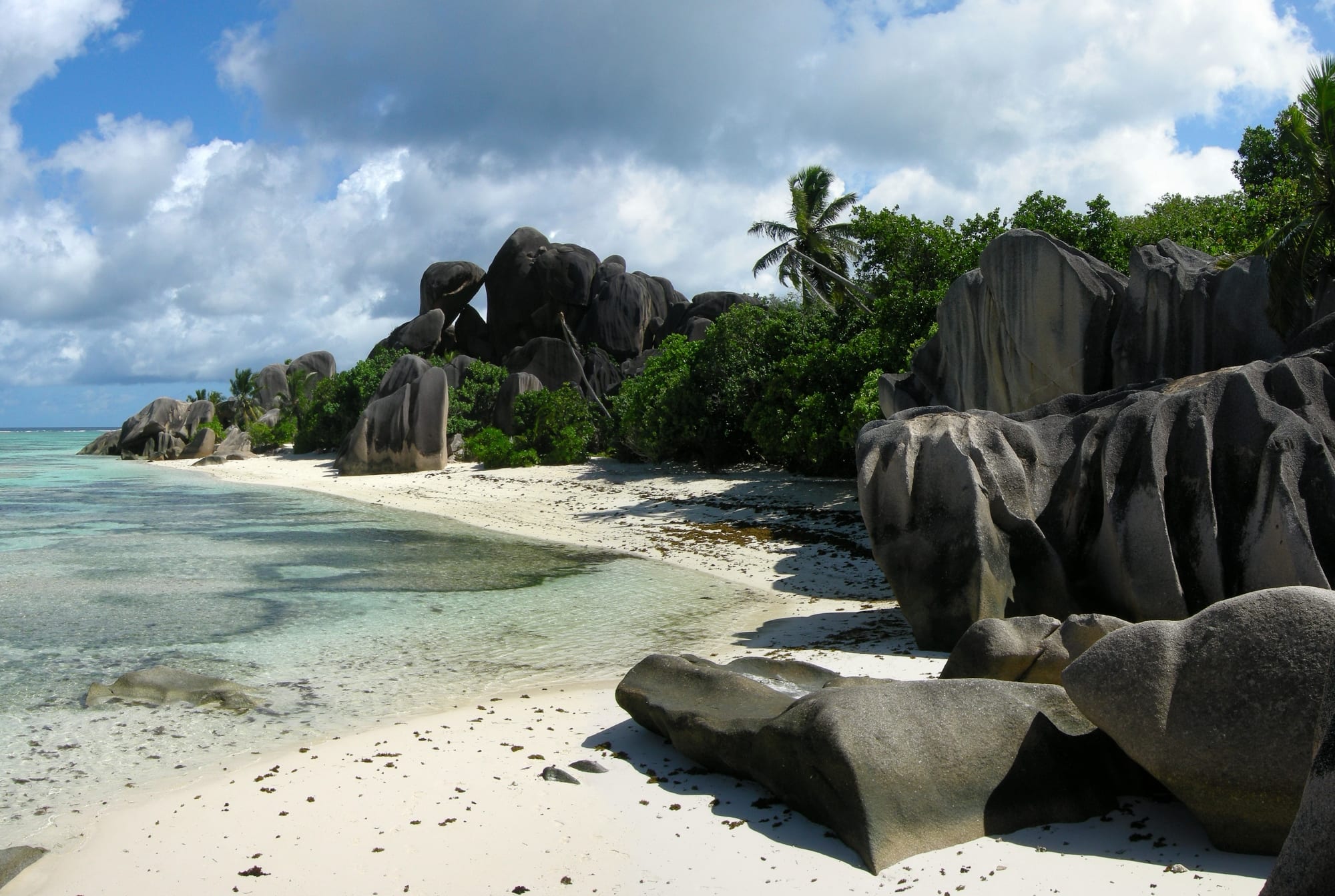 Huge smooth pink granite boulders on a white-sand beach beside pale turquoise shallows on La Digue