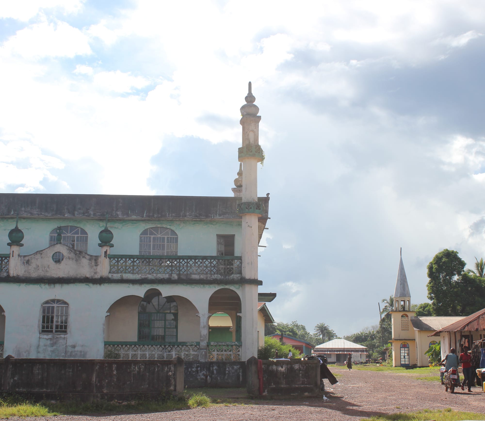 A two-storey pale green mosque with arched verandas and a tall white minaret on the left, and a small yellow church with a pointed steeple on the right, on a red dirt road under a cloudy sky