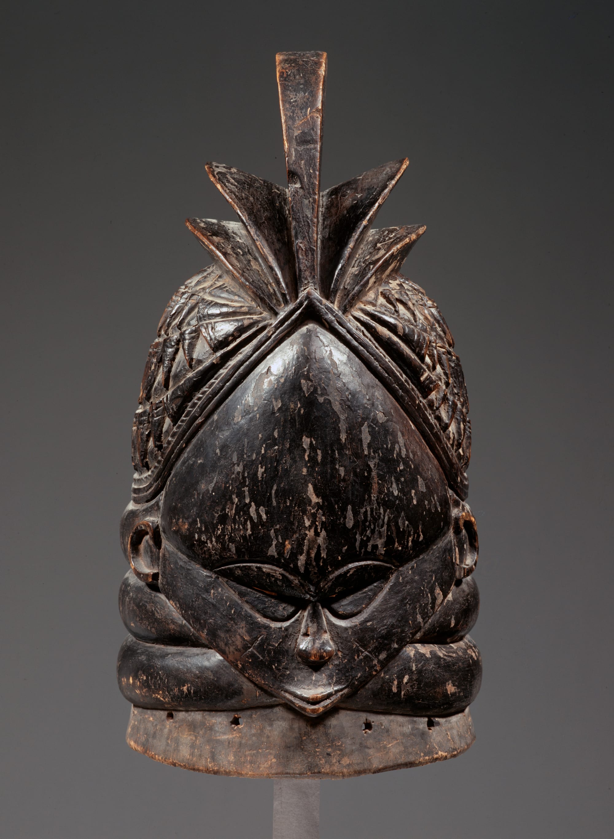 A carved black wooden helmet mask with a small face, closely set eyes, thick rings around the neck and a tall, crested hairstyle rising to a point