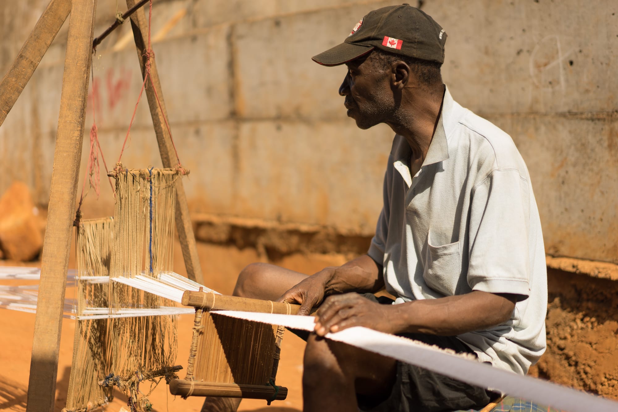 A man in a grey polo shirt and cap sits at a simple wooden loom, weaving a long narrow strip of white cotton cloth that stretches out in front of him