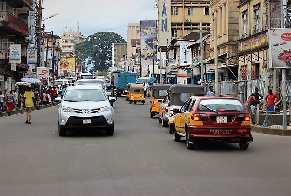 A busy Freetown street full of cars and minibus taxis, with a large cotton tree visible in the distance