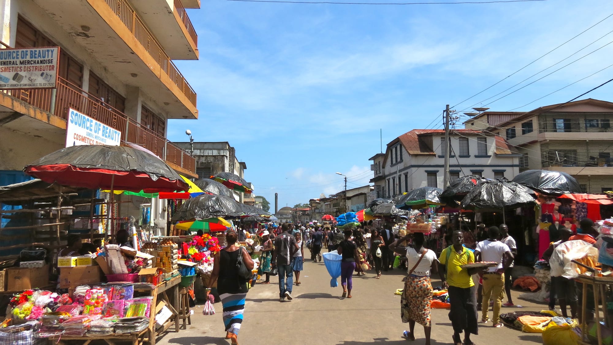 A crowded market street in Freetown lined with umbrella-covered stalls selling flowers, eggs and household goods