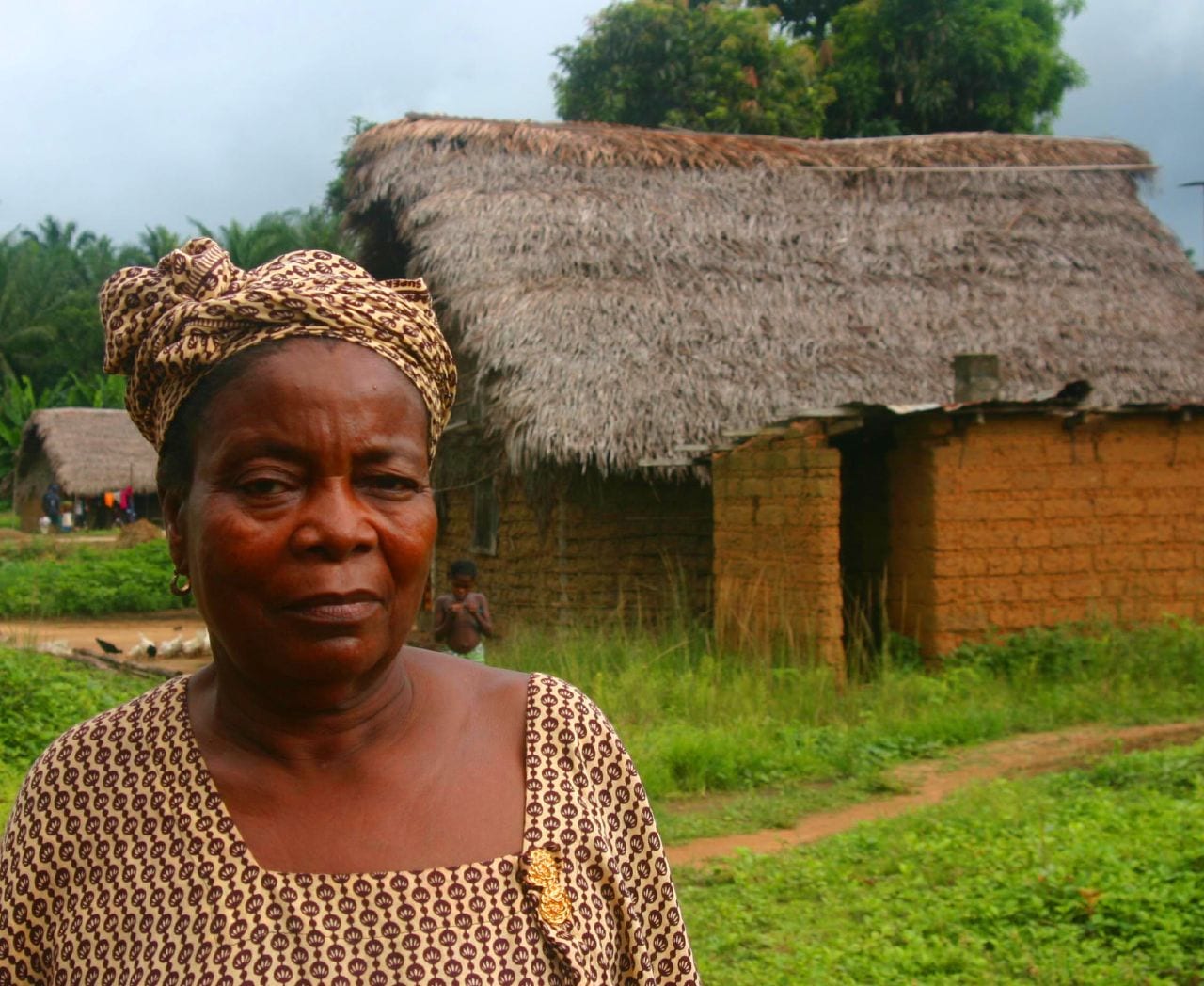 A woman in a patterned headwrap and dress standing in front of a thatched-roof mud-brick house, with a child visible behind her