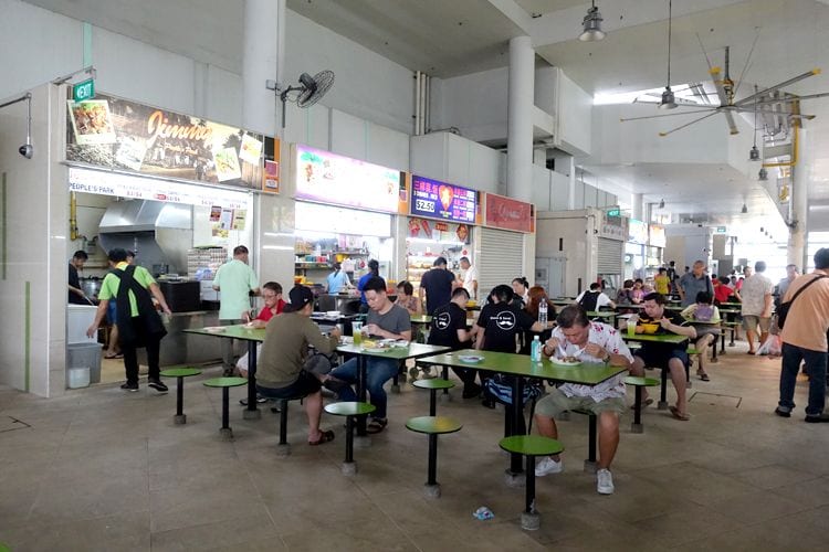 People sitting at tables eating and queuing at food stalls inside Bedok Hawker Centre