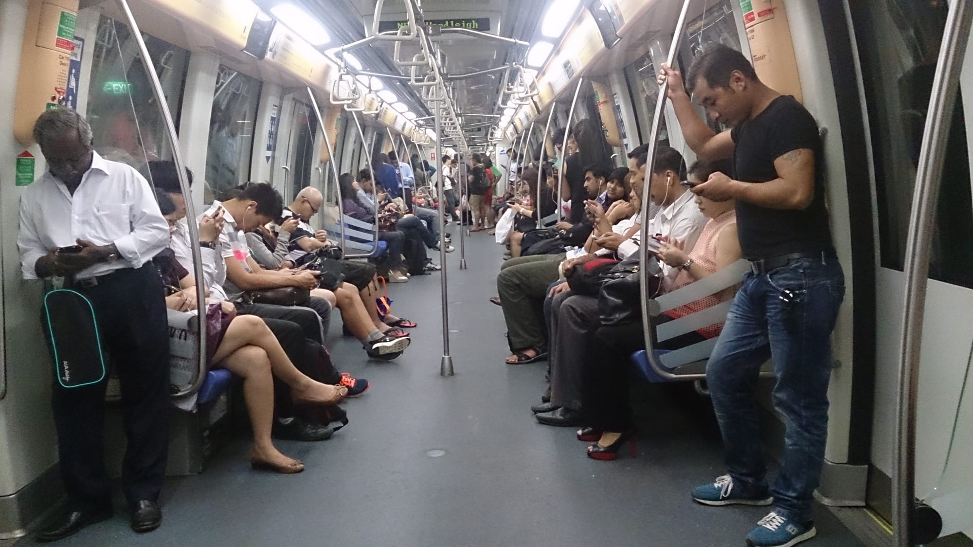 Commuters standing and sitting inside a Singapore MRT train carriage, most looking at their phones