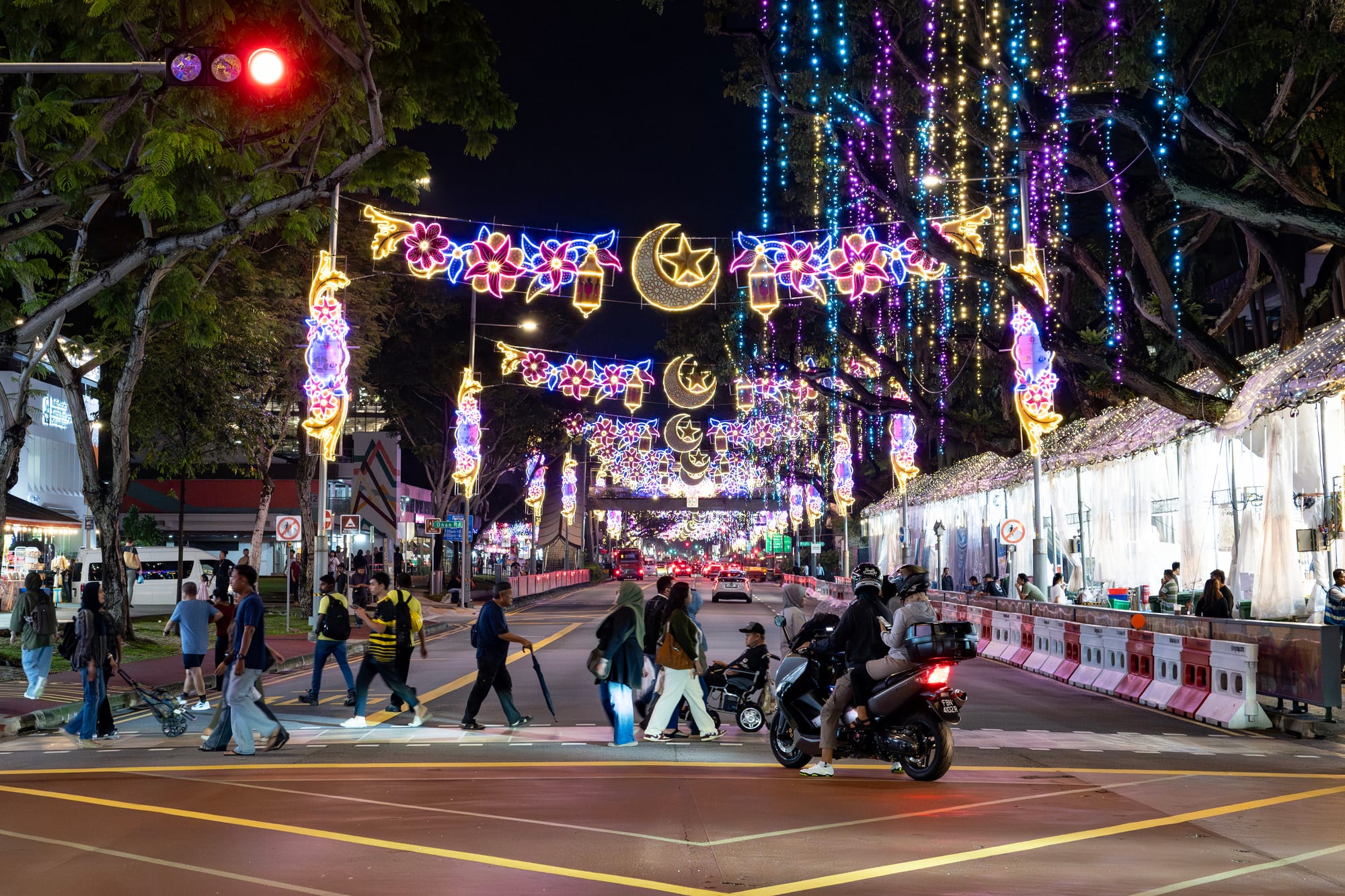 A wide street at night strung with illuminated decorations of crescent moons, stars, flowers and lanterns, with pedestrians crossing in the foreground, some women in headscarves