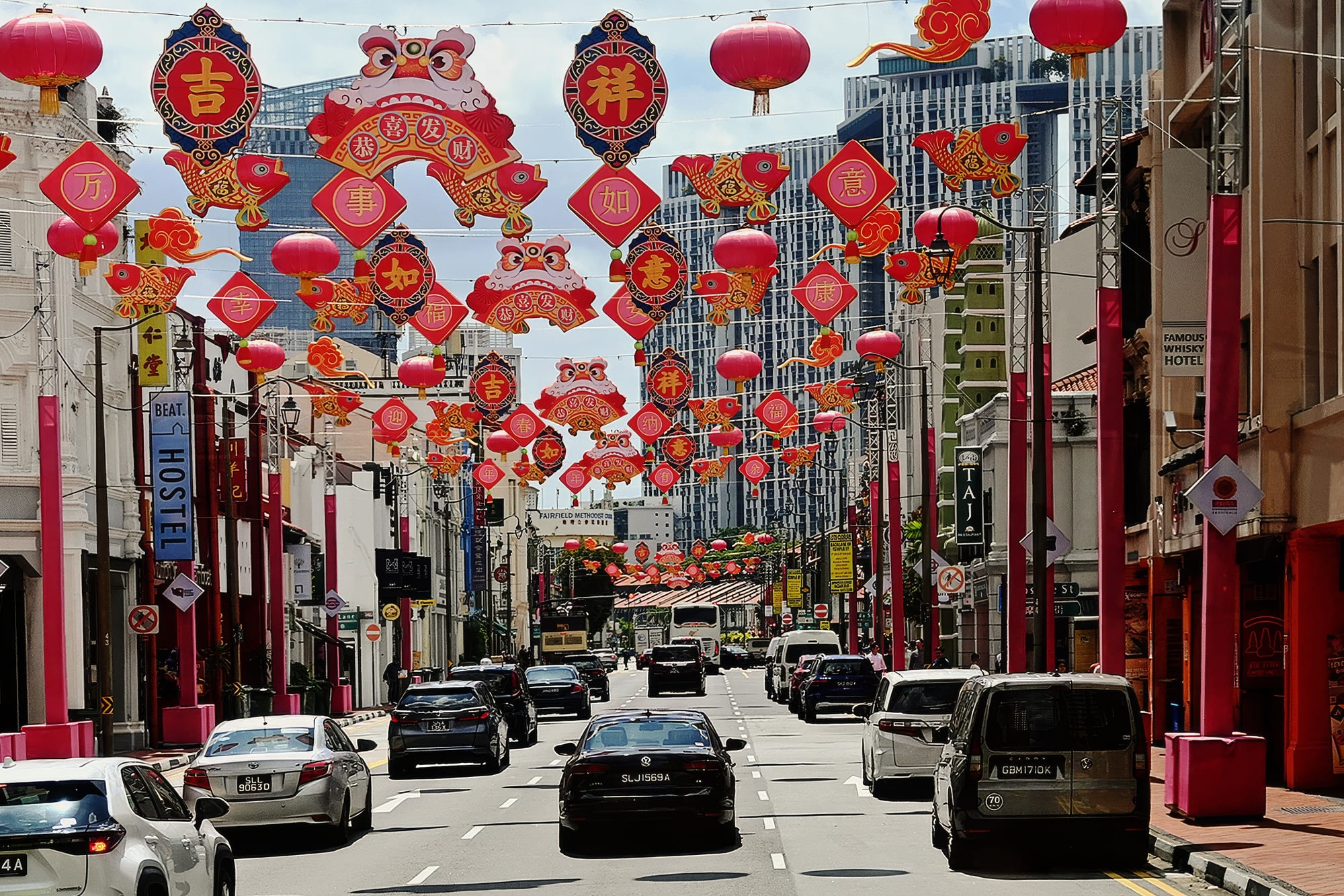 A city street lined with shophouses and tall office towers, hung overhead with red lanterns, red diamond-shaped signs bearing Chinese characters, lion heads and golden fish, with cars driving below