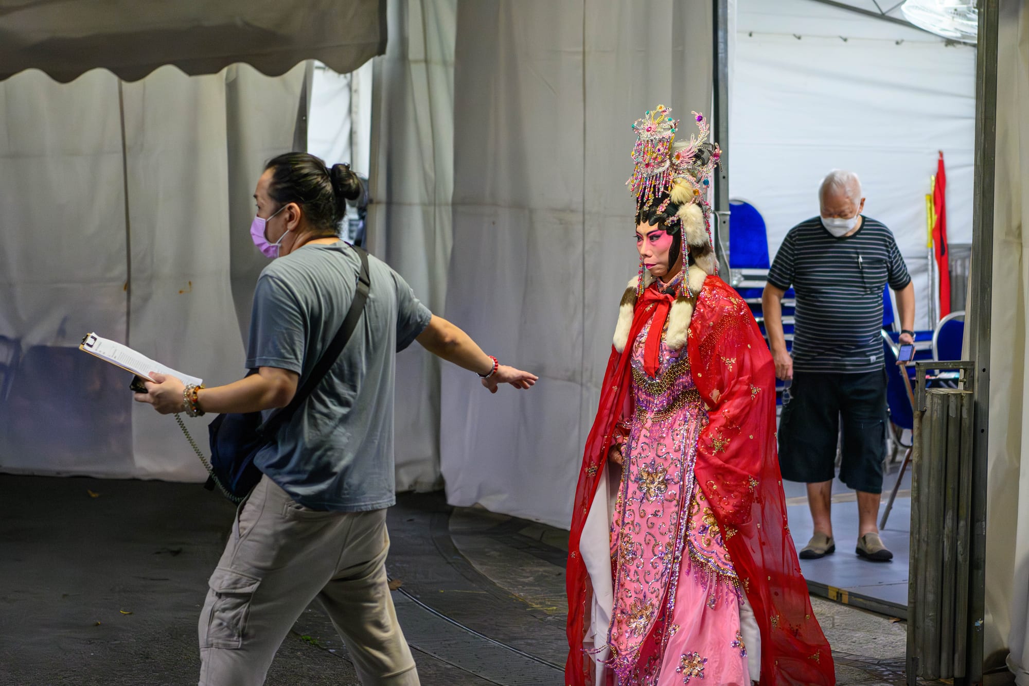 A Chinese opera performer in an ornate jewelled headdress, pink embroidered costume and flowing red cape walks through a backstage tent, with a crew member gesturing ahead and an older man watching behind