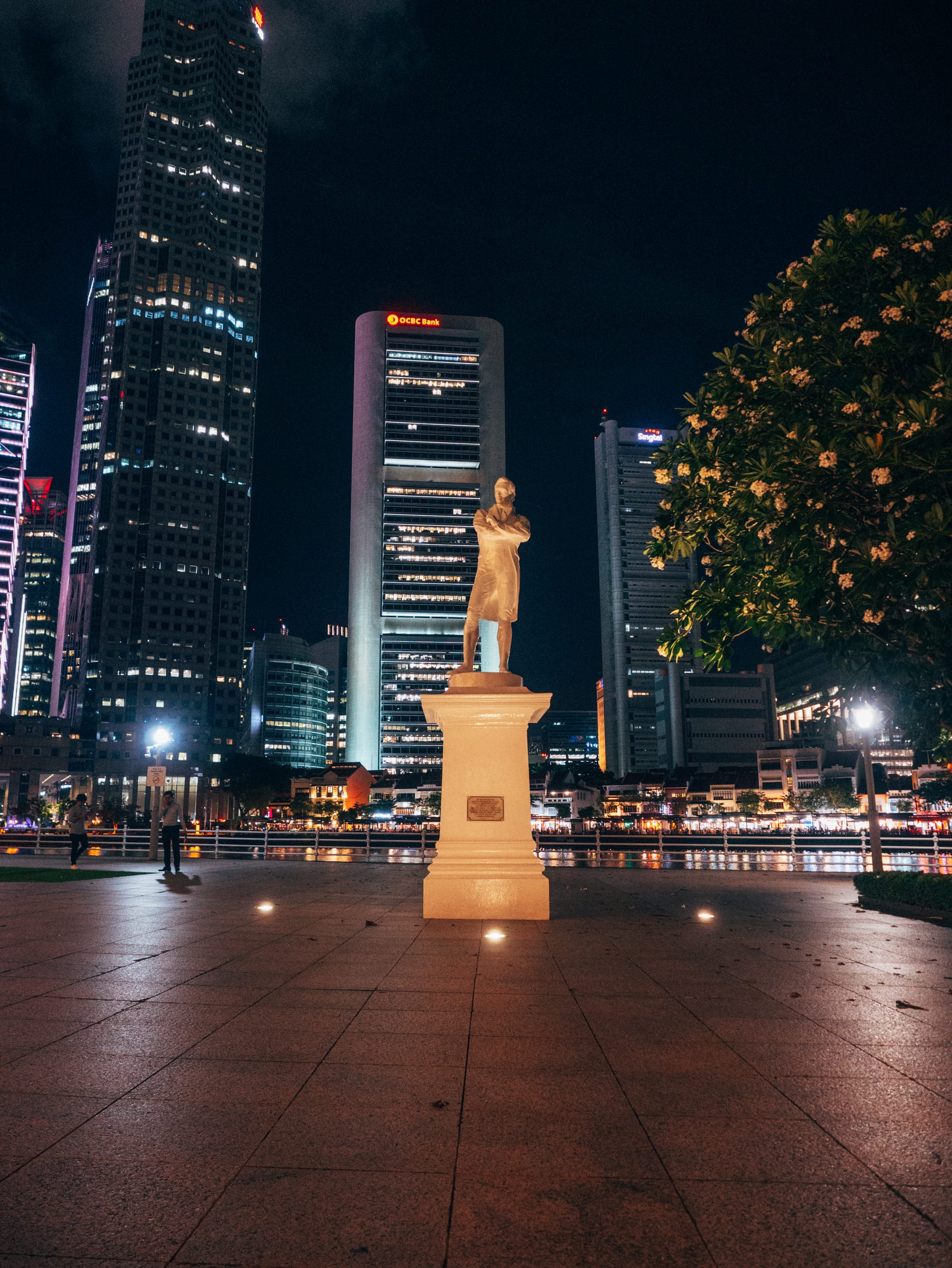 A white statue of Stamford Raffles standing with arms folded on a plinth by the Singapore River