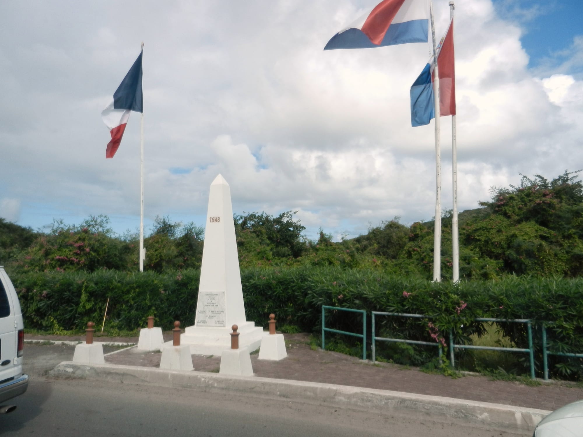 A white stone obelisk marked 1648 stands on a small paved area beside a road, flanked by a French flag on one side and the Dutch and Sint Maarten flags on the other, with green hills behind