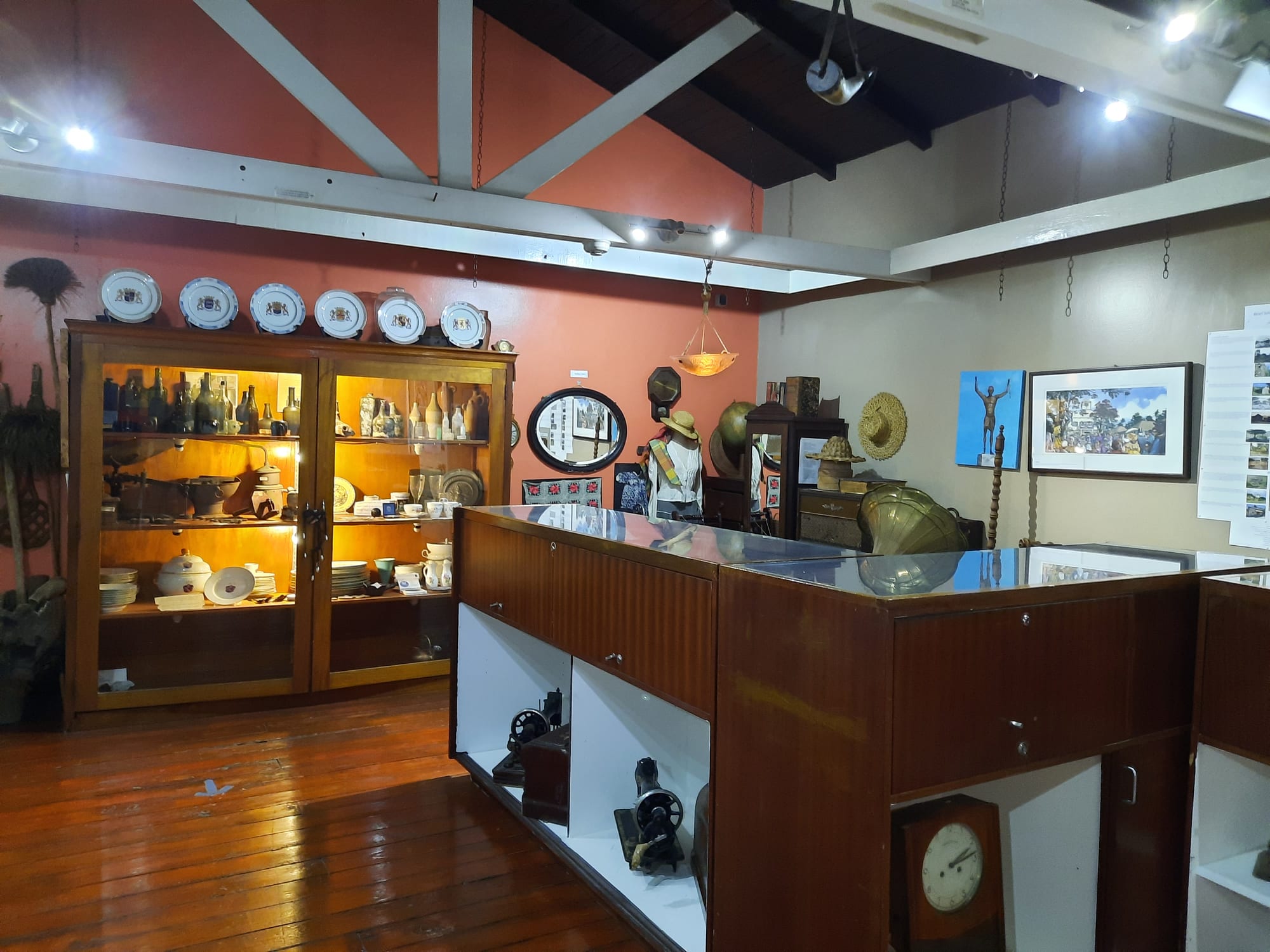 A museum room with a wooden display cabinet of old bottles, jugs and china, decorated plates along the top, straw hats, a mirror and old sewing machines on shelves beneath a glass counter