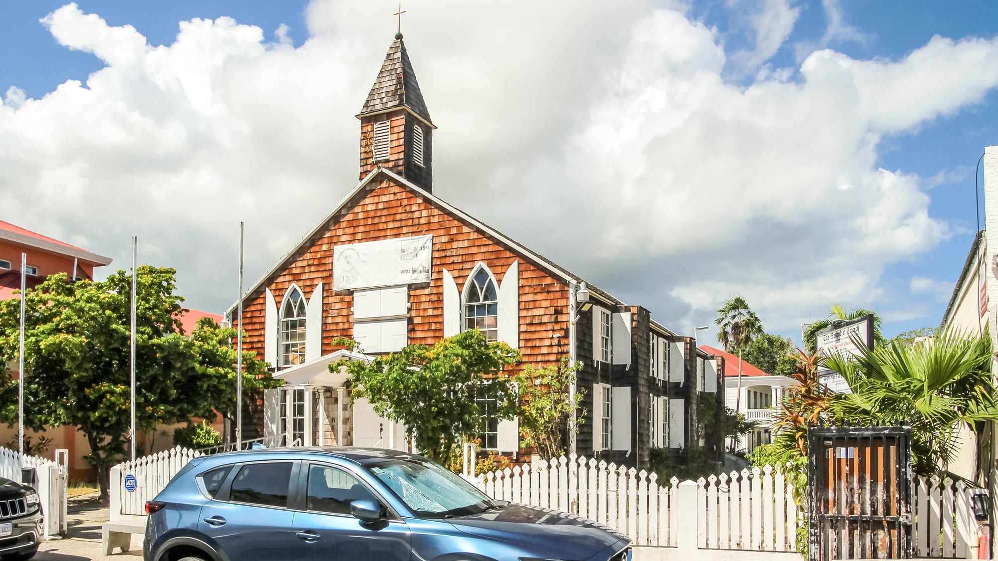 A small church clad in brown wooden shingles with a pointed bell tower, arched white-shuttered windows and a white picket fence, under a bright sky with tall clouds