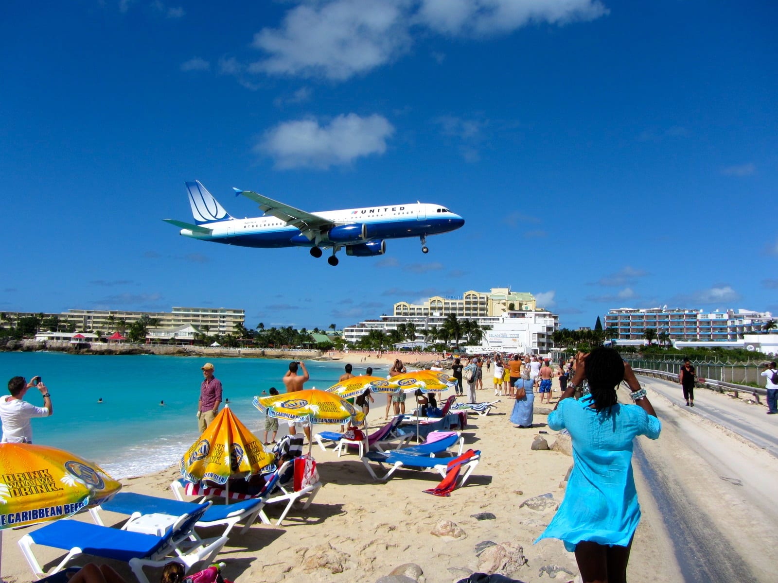 A low-flying aircraft passing directly over a beach and a road just before an airport runway