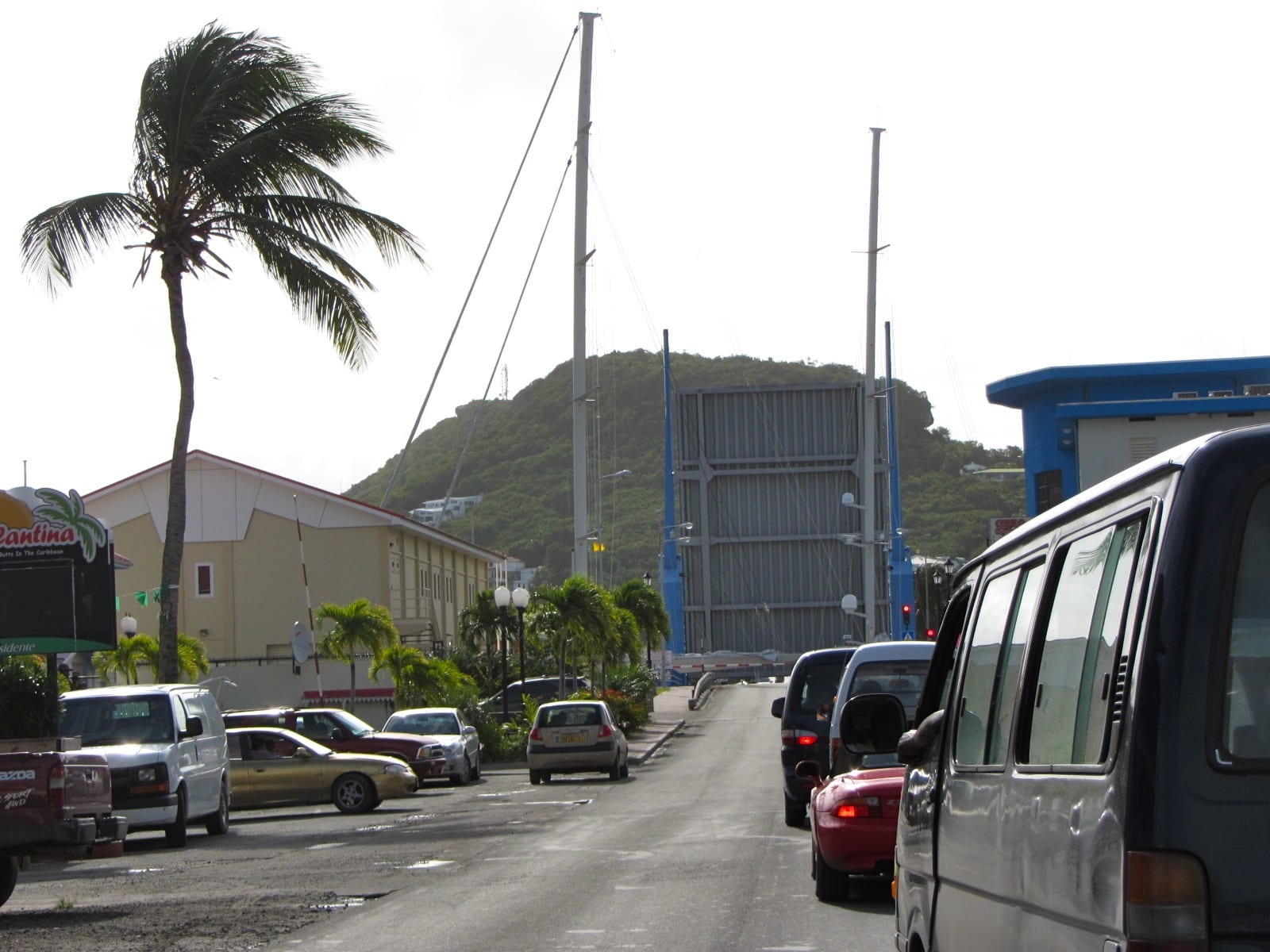 Cars queued on a road as the Simpson Bay drawbridge stands raised in the background, with a palm tree in the foreground
