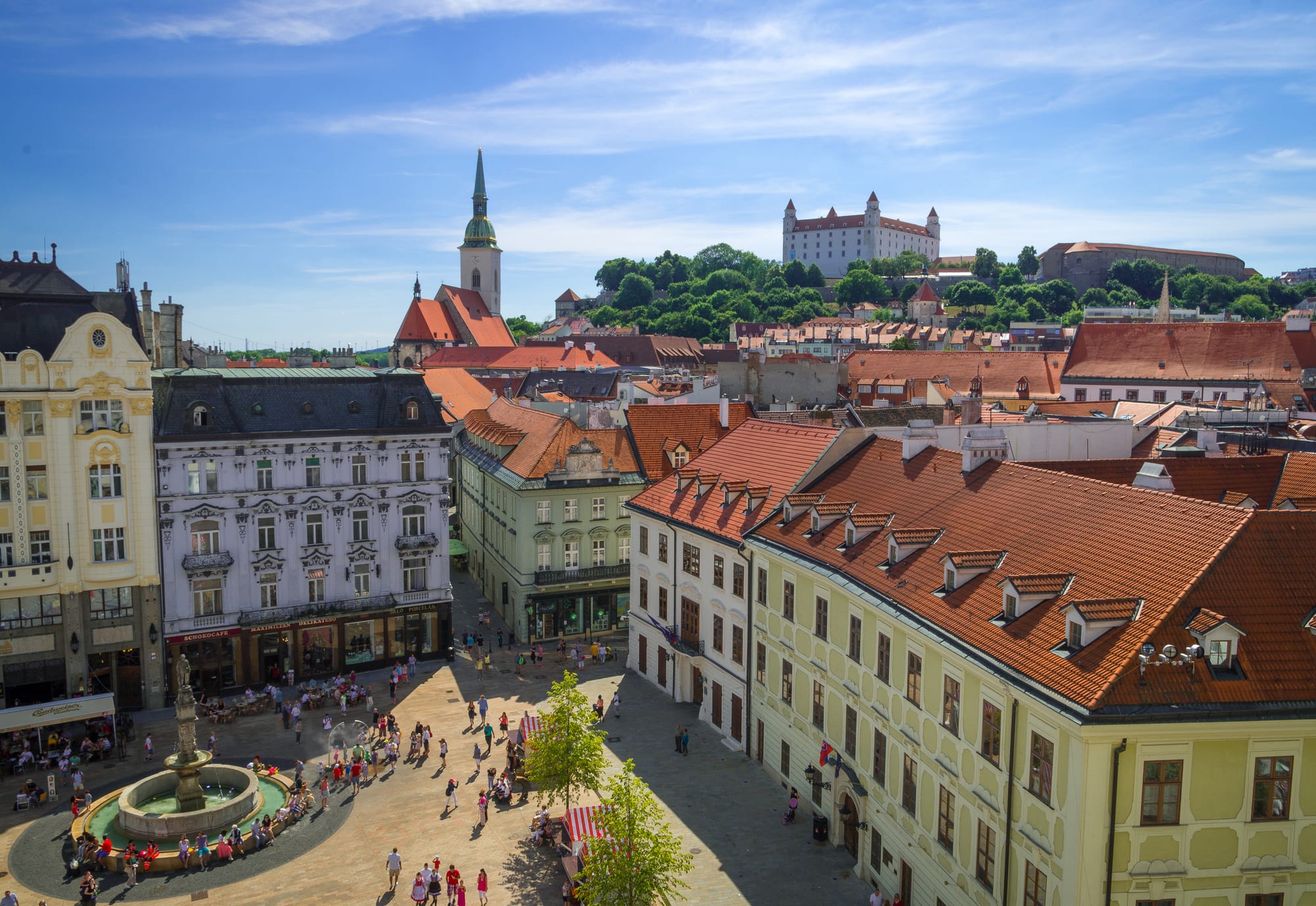 An aerial view of a historic town square with a fountain, café tables and people walking, a castle visible on a hill behind