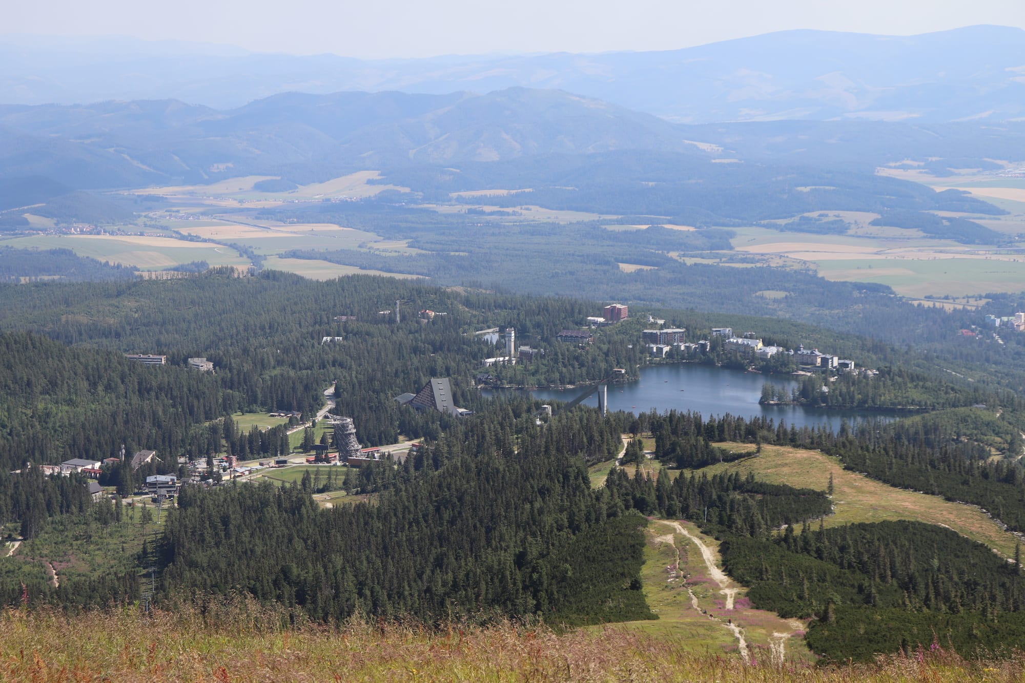 An aerial view over a forested mountain resort with a small lake, hotels and a hiking trail, with hazy peaks in the distance