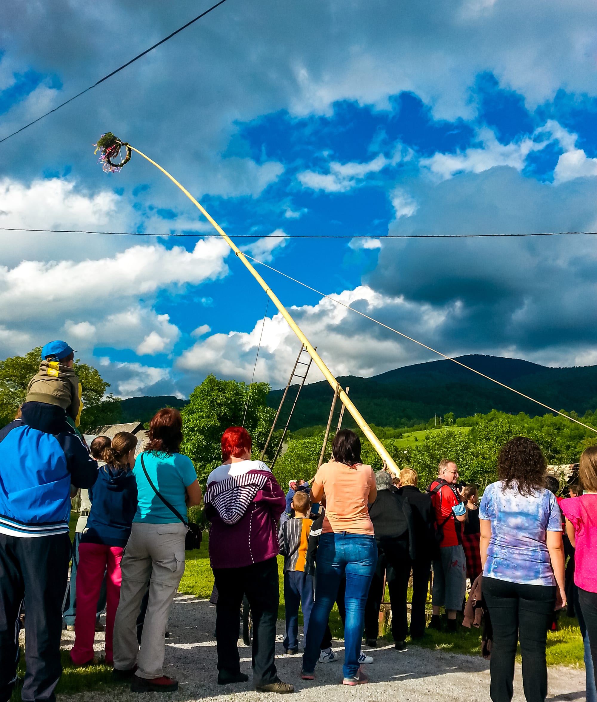 Villagers stand with their backs to the camera watching a tall, pale maypole, topped with a small wreath of greenery and ribbons, being hauled upright with ropes and ladders against a cloudy blue sky and green hills