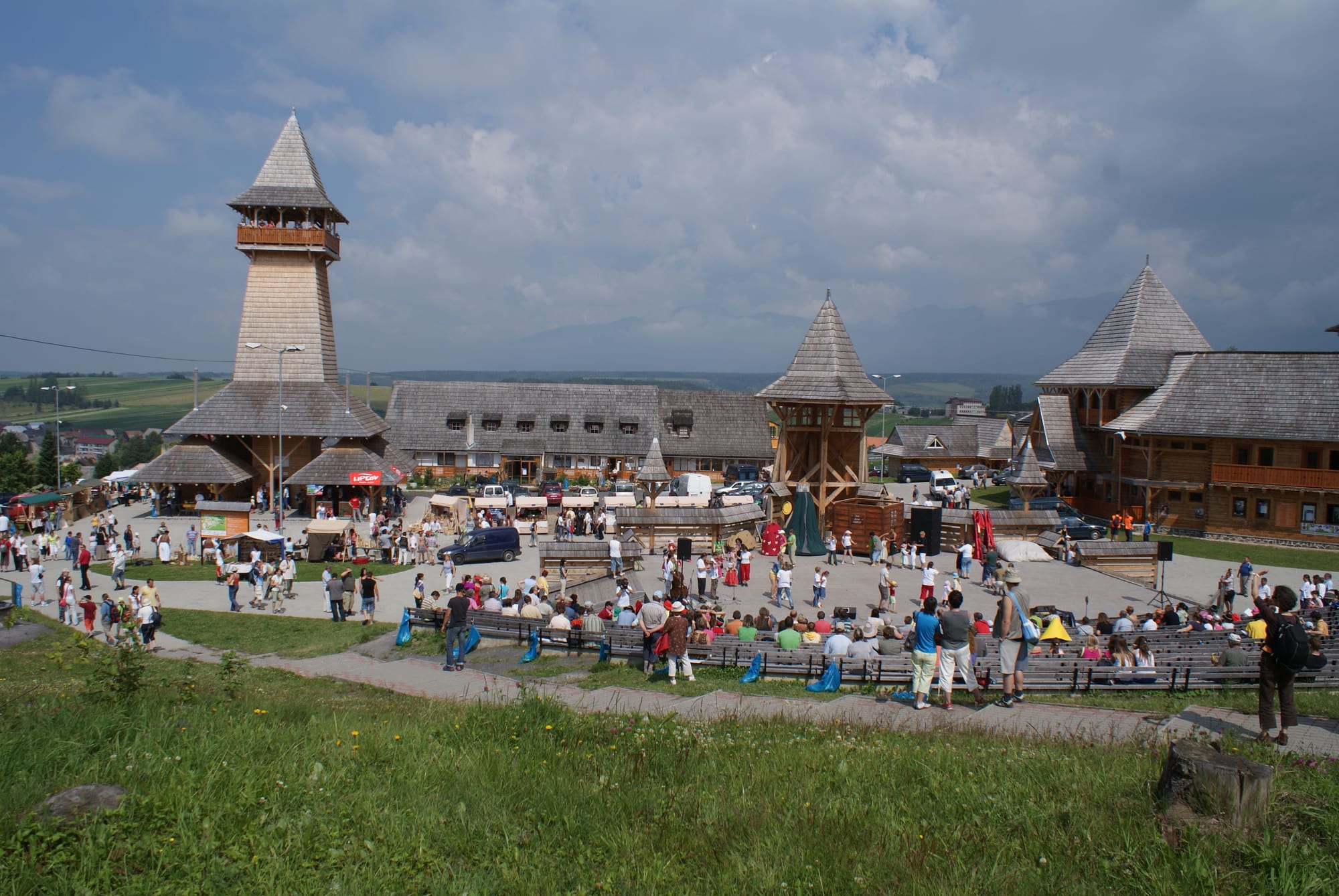 A crowd gathered on benches around an open-air stage, surrounded by wooden buildings with steep shingled roofs and a tall shingled tower, with fields and hills beyond under a cloudy sky