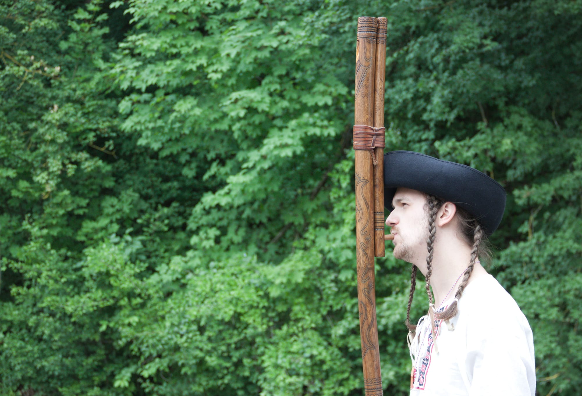 A young man with long braids, a wide black hat and a white embroidered shirt, seen in profile, plays a very long, carved wooden flute held vertically, against a background of green trees