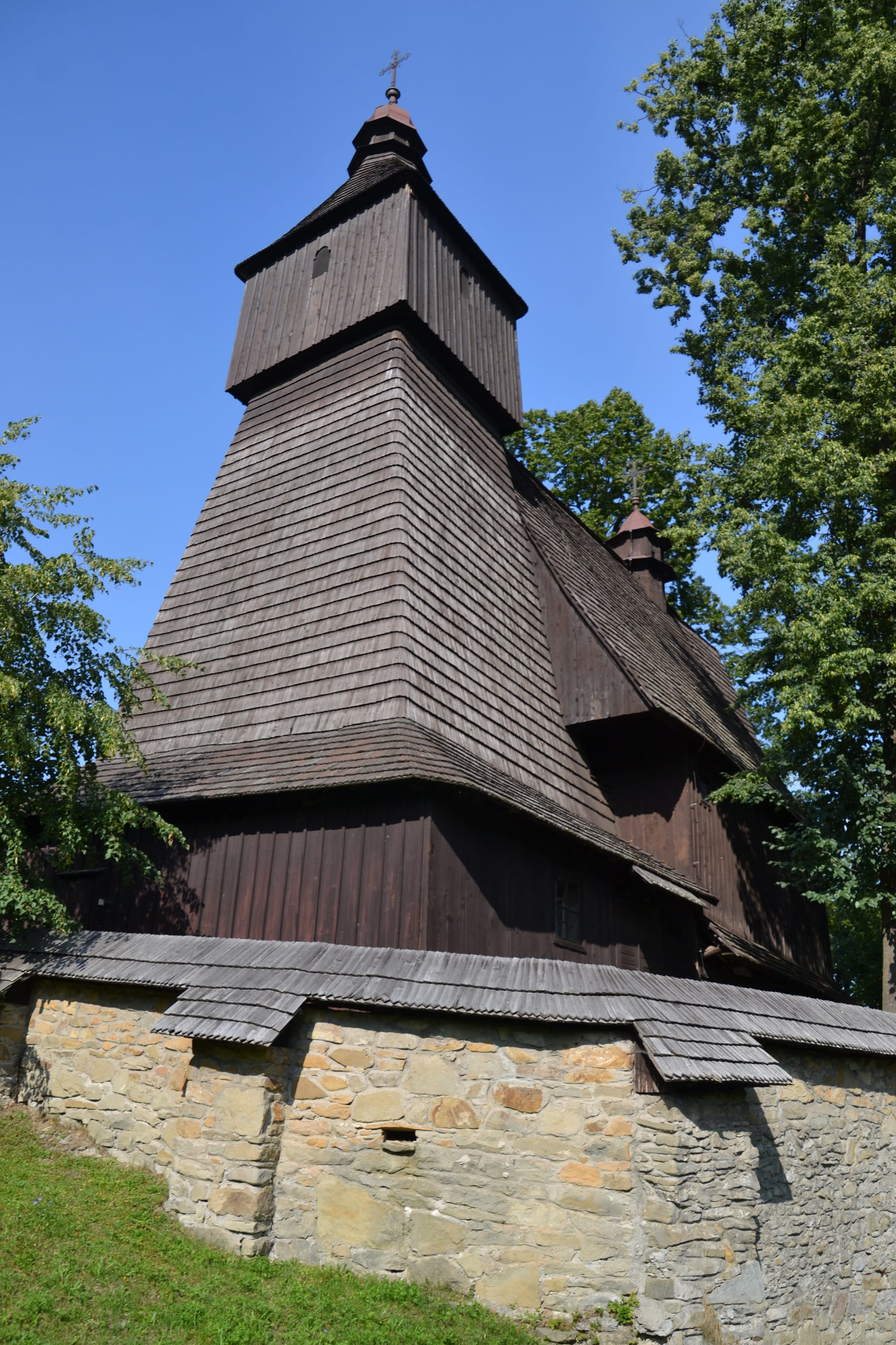A dark wooden church with a tall shingled bell tower topped by a small onion-shaped cupola and cross, rising above a low stone wall with a shingled coping, framed by trees against a blue sky