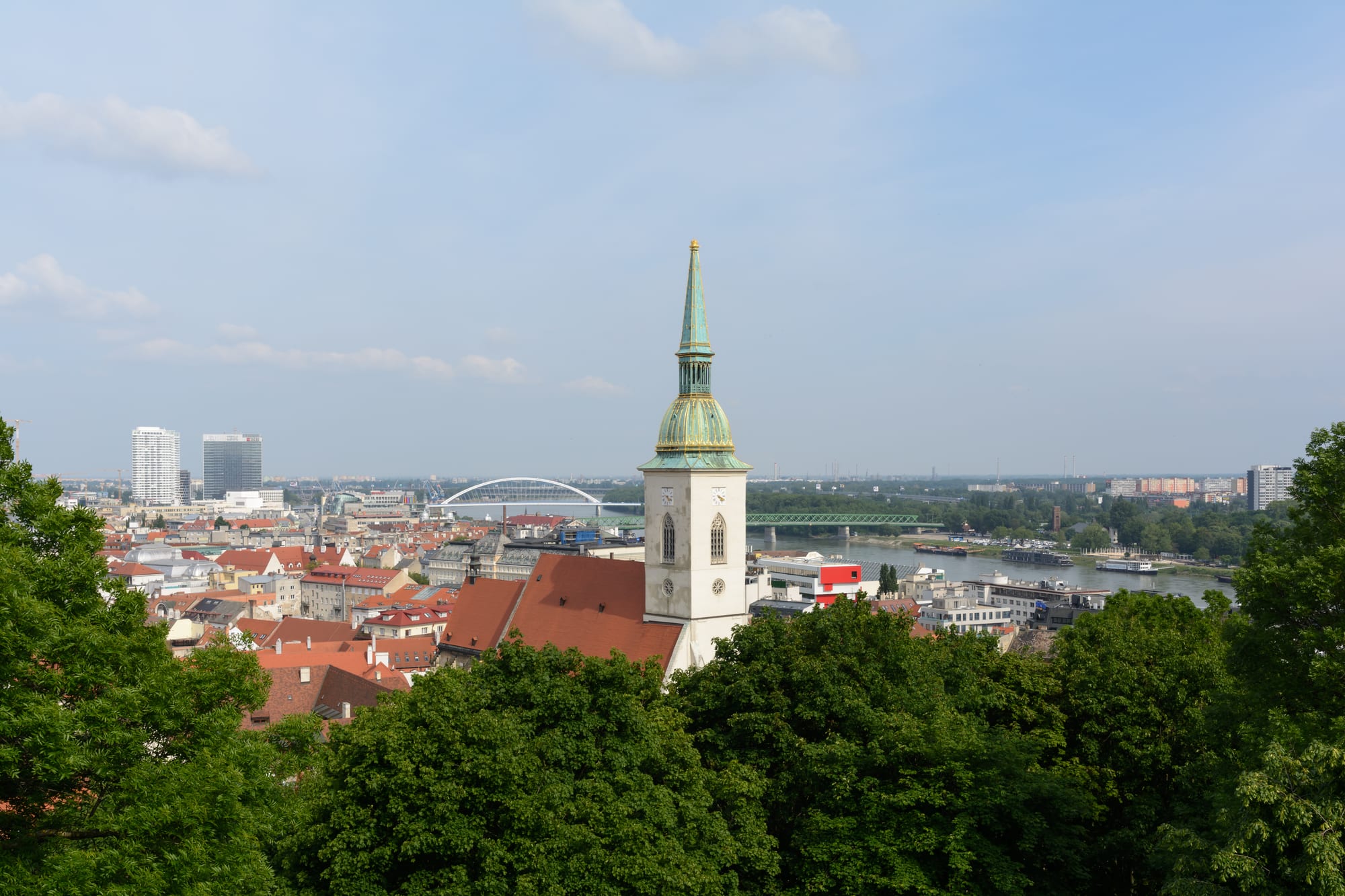 The white tower and green copper spire of Saint Martin's Cathedral rising above the red roofs of Bratislava, with the Danube and its bridges behind