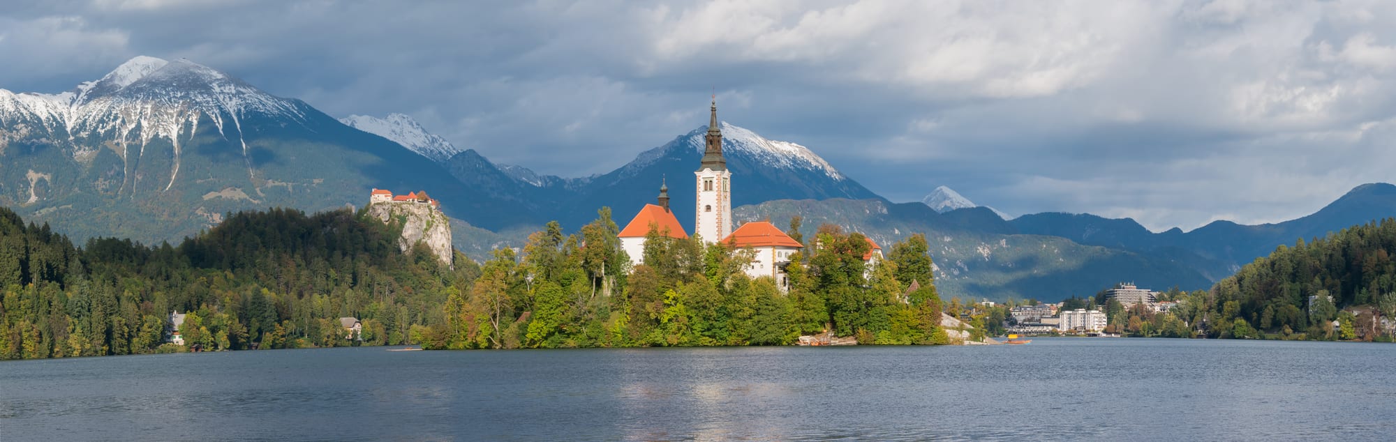A small church on a green island in the middle of an alpine lake, with a castle on a cliff above
