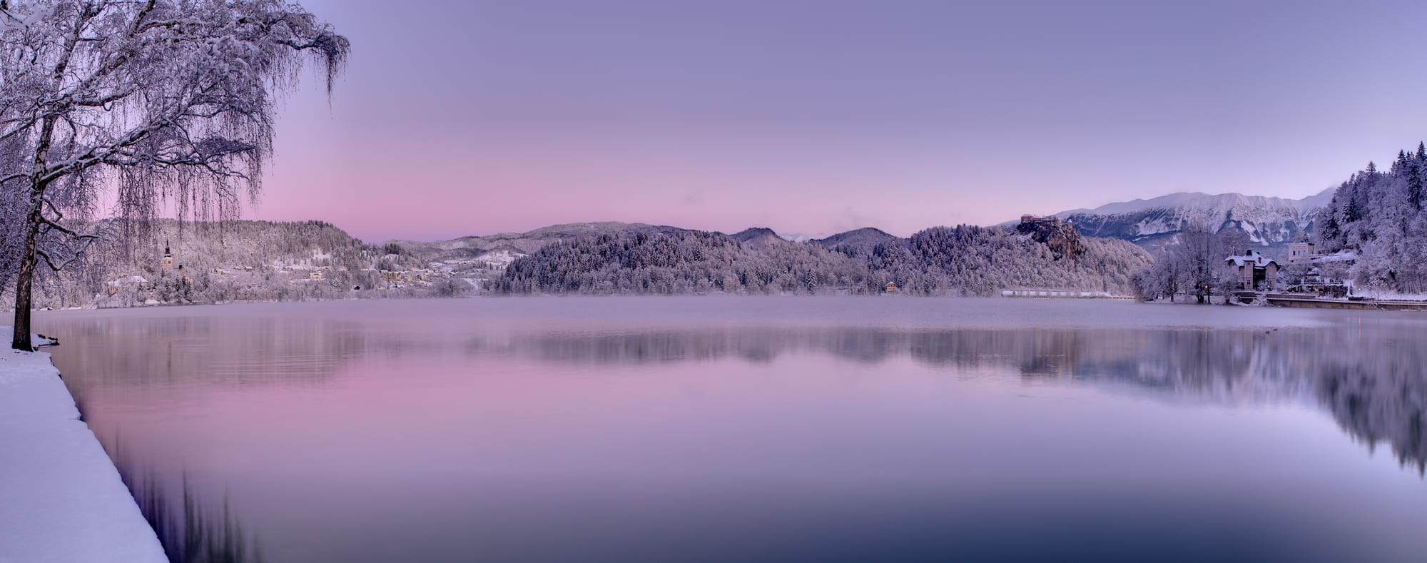 A small church on an island in an alpine lake, with a clifftop castle and snow-dusted mountains beyond