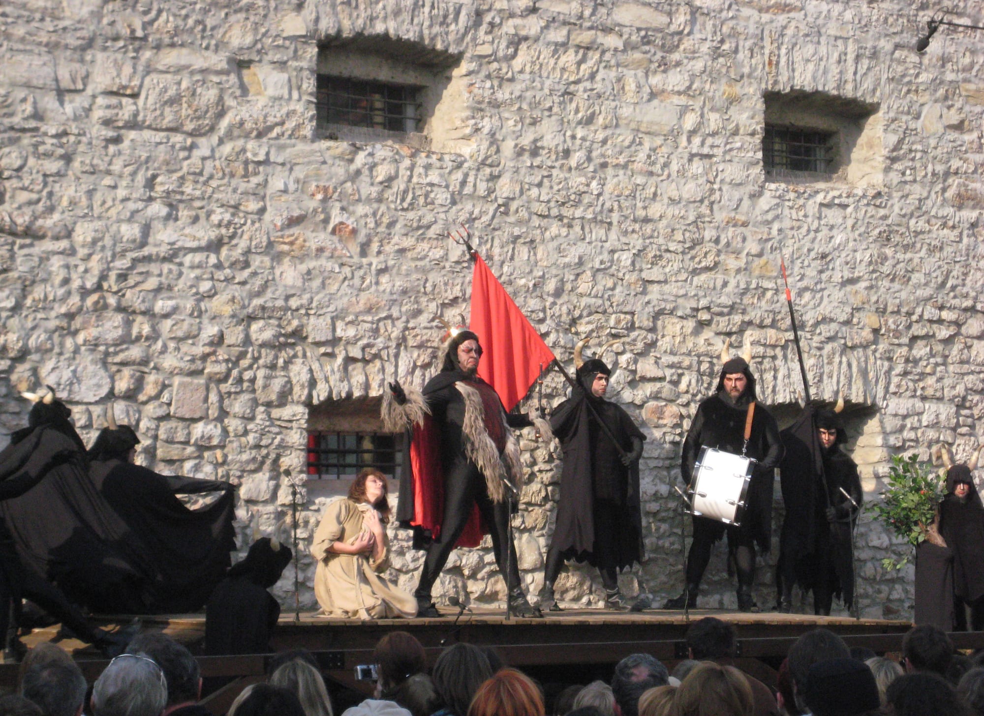 Actors in black hooded costumes with horns perform on an outdoor stage before a stone wall, one waving a red flag, another beating a drum, while a figure in a pale robe kneels among them and an audience watches below