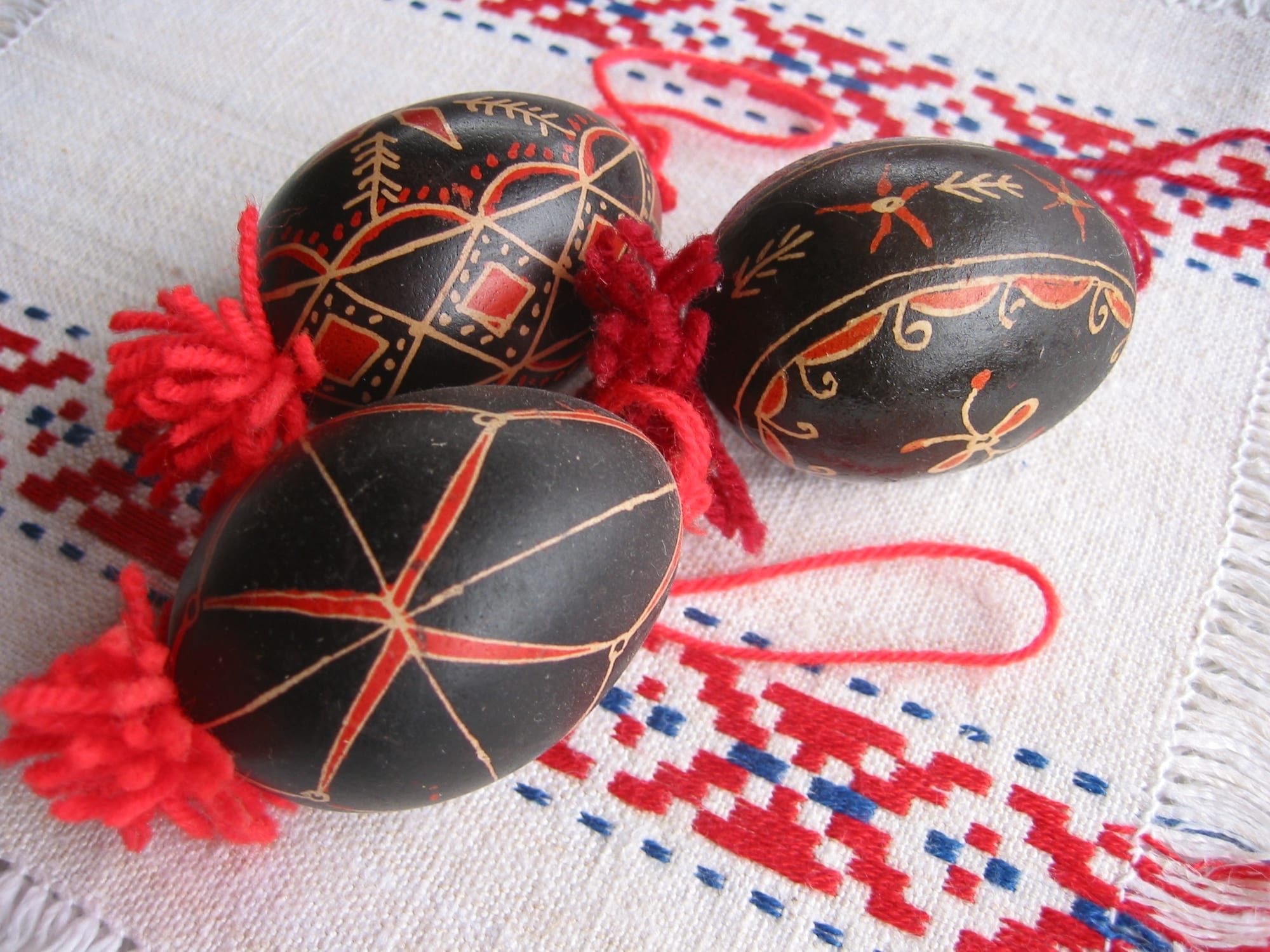 Three dark Easter eggs decorated with fine red and pale lines, stars and geometric patterns, each with a red wool tassel, lying on a white cloth embroidered in red and blue