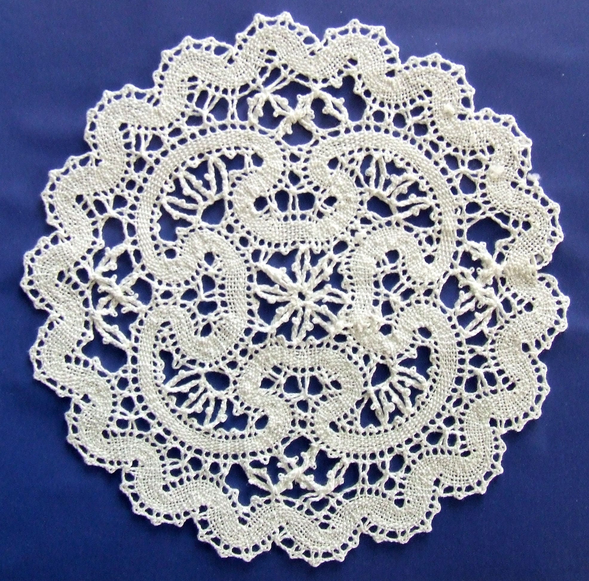 A round white bobbin-lace doily with wavy bands, flower-like motifs and a scalloped edge, laid on a deep blue background