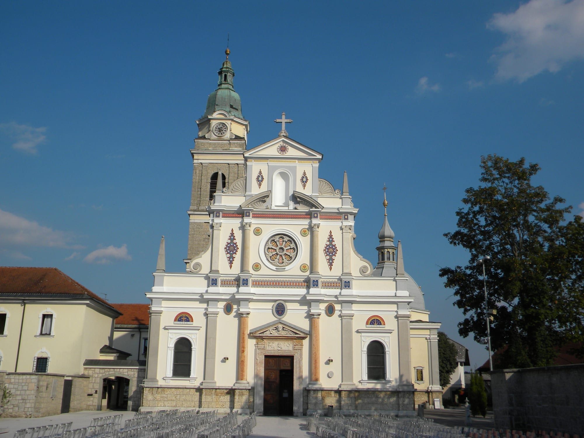 The pale yellow and white facade of a large church with a rose window, colourful mosaic decoration and a green-domed bell tower, with rows of empty chairs set out in the square in front under a blue sky