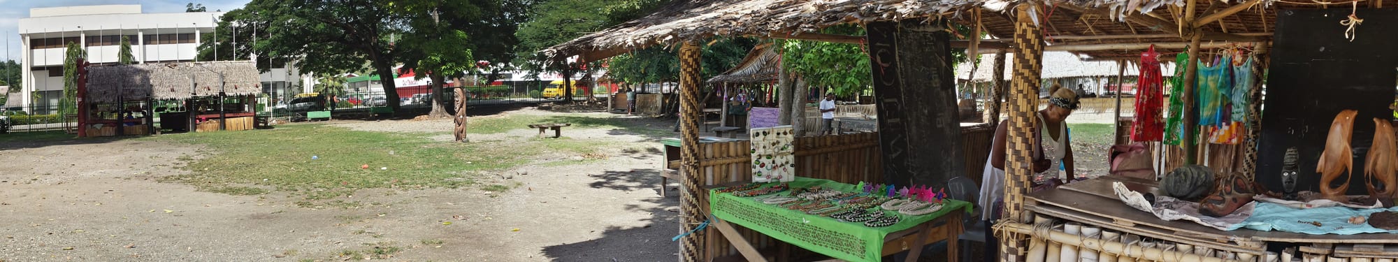 Open-sided thatched market stalls displaying carved wooden items and jewellery, with a vendor arranging goods