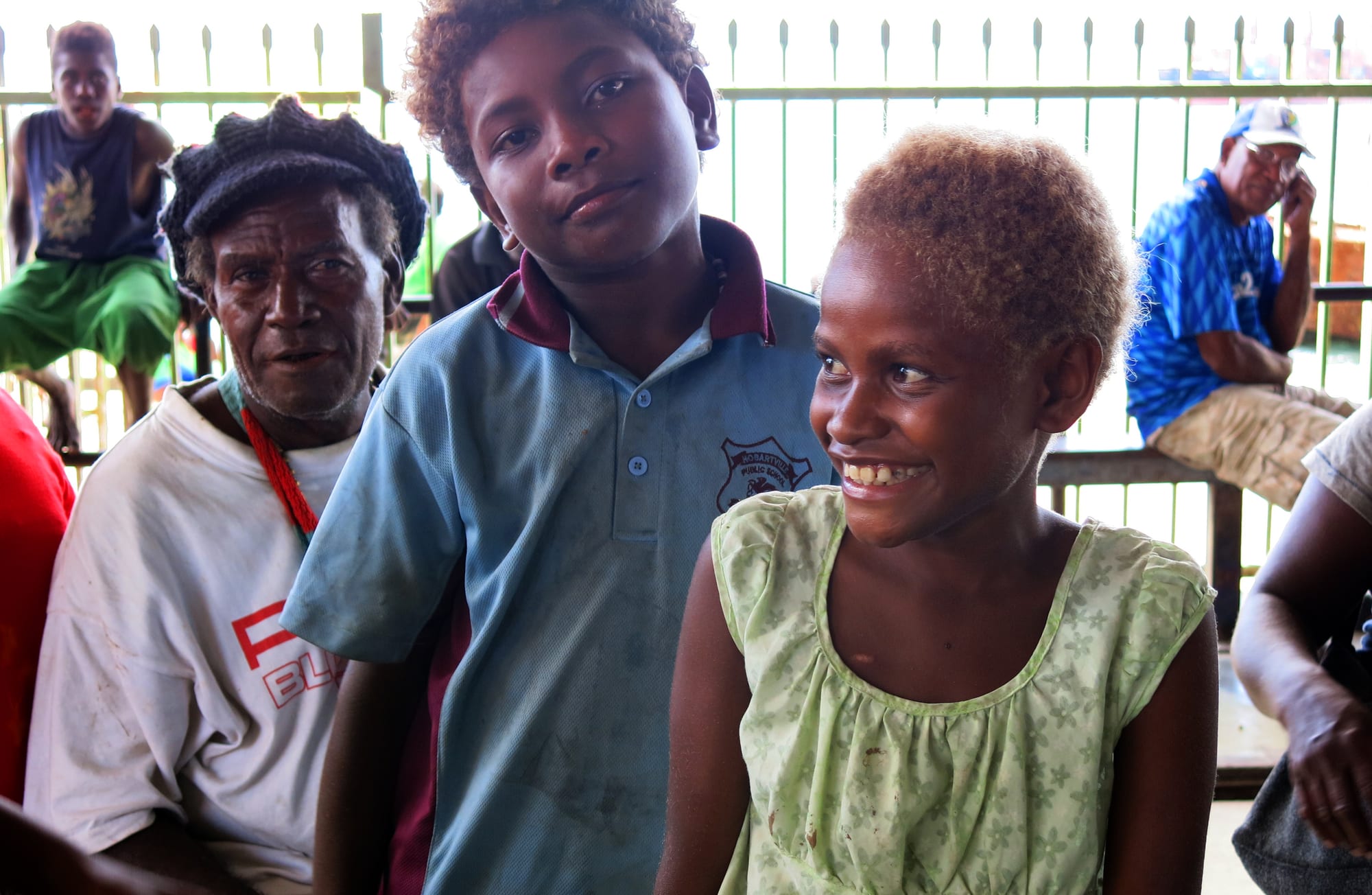 An older man and two smiling children sitting together at a market, with other people visible behind a railing