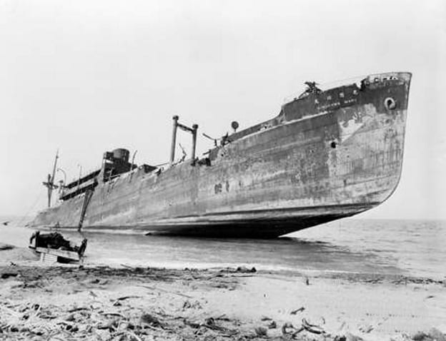A black-and-white photo of a large rusting cargo ship run aground on a beach