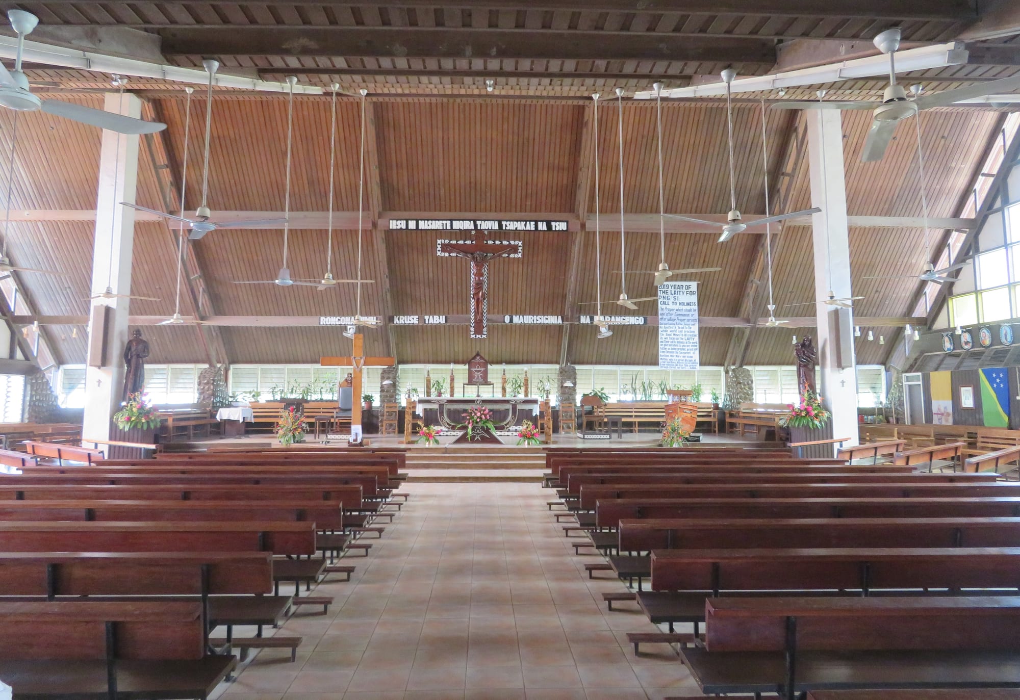 The interior of a large church with a high wooden ceiling, rows of ceiling fans, long wooden pews on a tiled floor, a crucifix and banners above the altar, flowers around the sanctuary and the national flag on the right